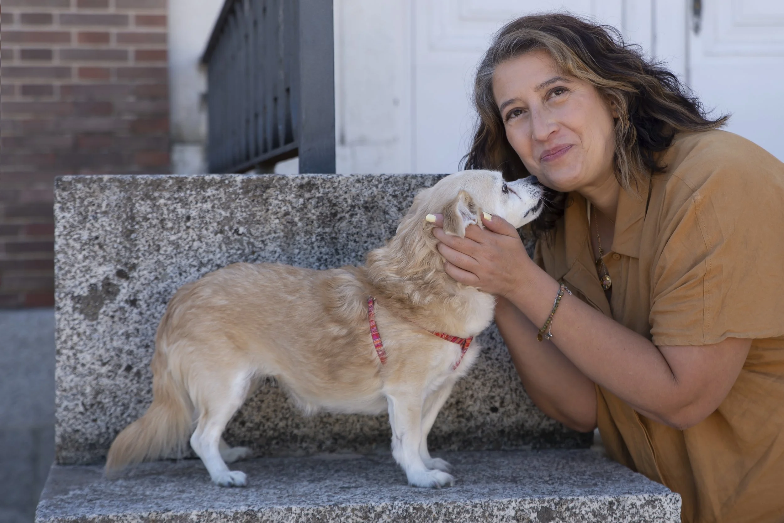Una mujer acariciando a un perro pequeño de color dorado sentado en una banca de piedra.