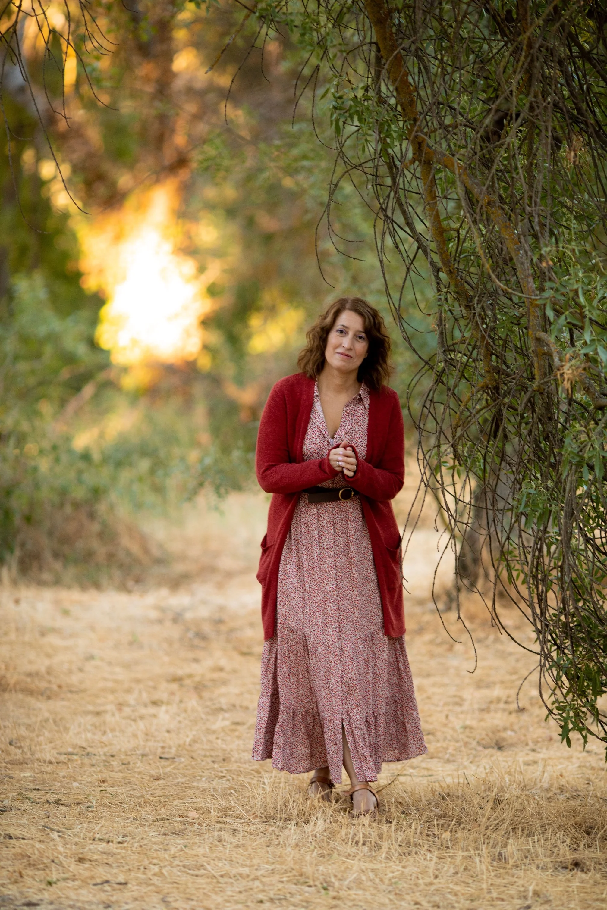 Una mujer con cabello rizado y expresión pensativa en un sendero rodeado de árboles y vegetación, con luz de atardecer en el fondo.