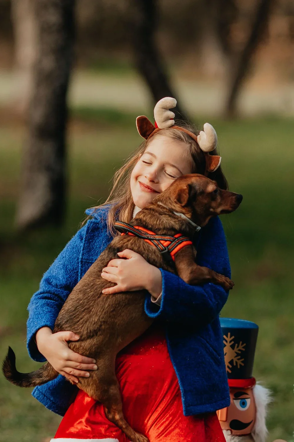 Niña sonriendo con orejas de reno abrazando a un perro marrón en un parque