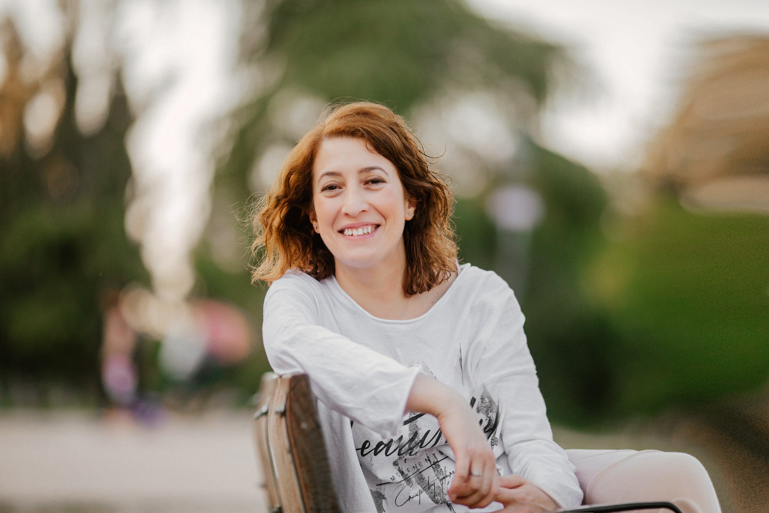 Mujer con cabello rizado y rubio sonriendo y sentado en un banco en un parque, con fondo difuminado de árboles y césped.