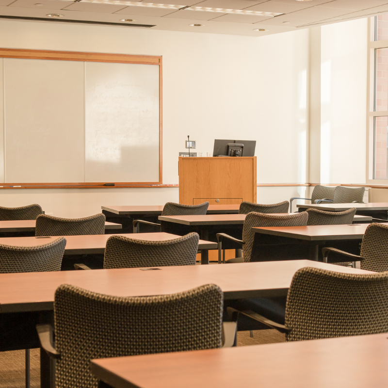 Aula  con escritorios y sillas, pizarra blanca y escritorio del profesor con computadora.