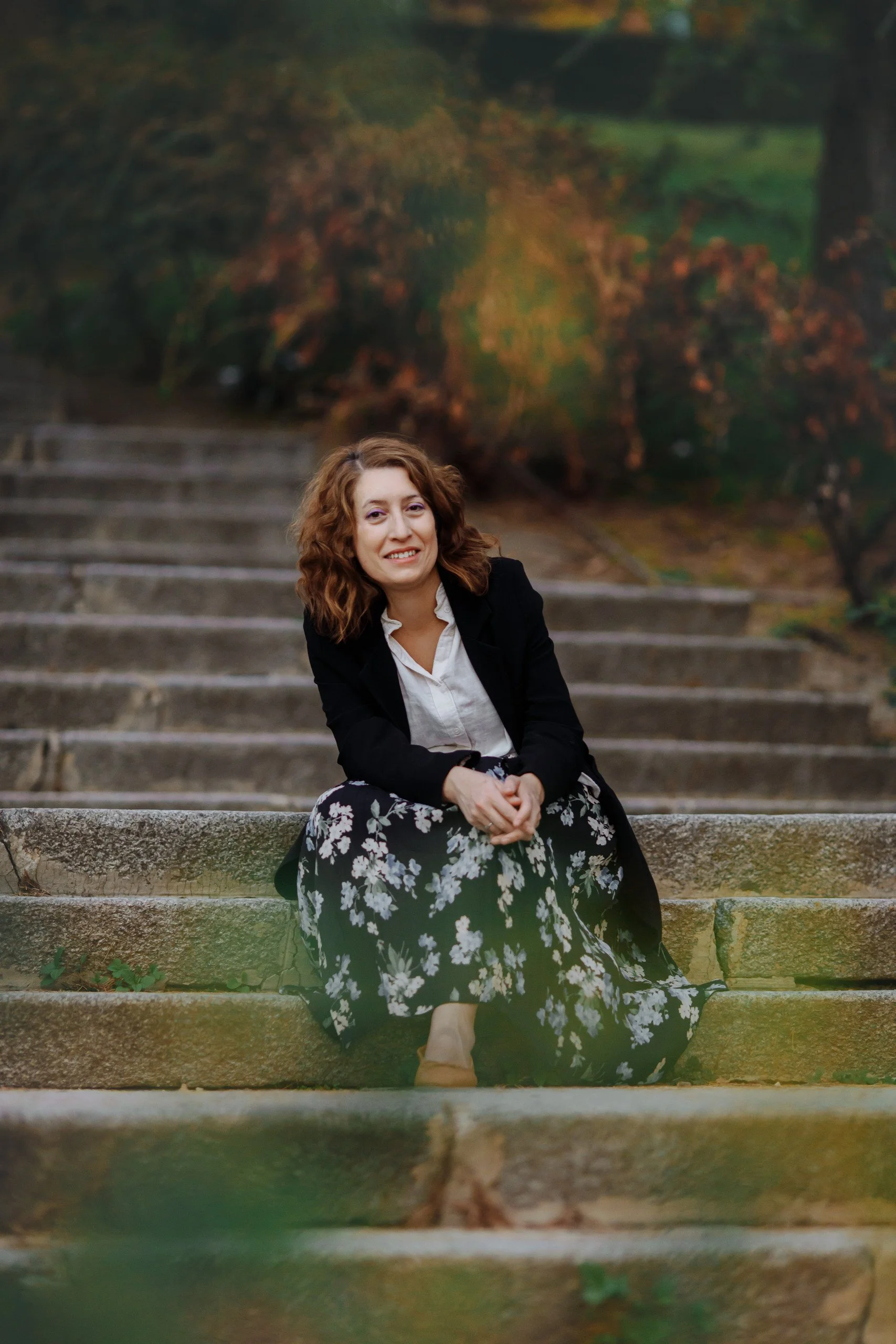 Profesora de Economía y Empresa sentada en escaleras de piedra en un parque con árboles de fondo.