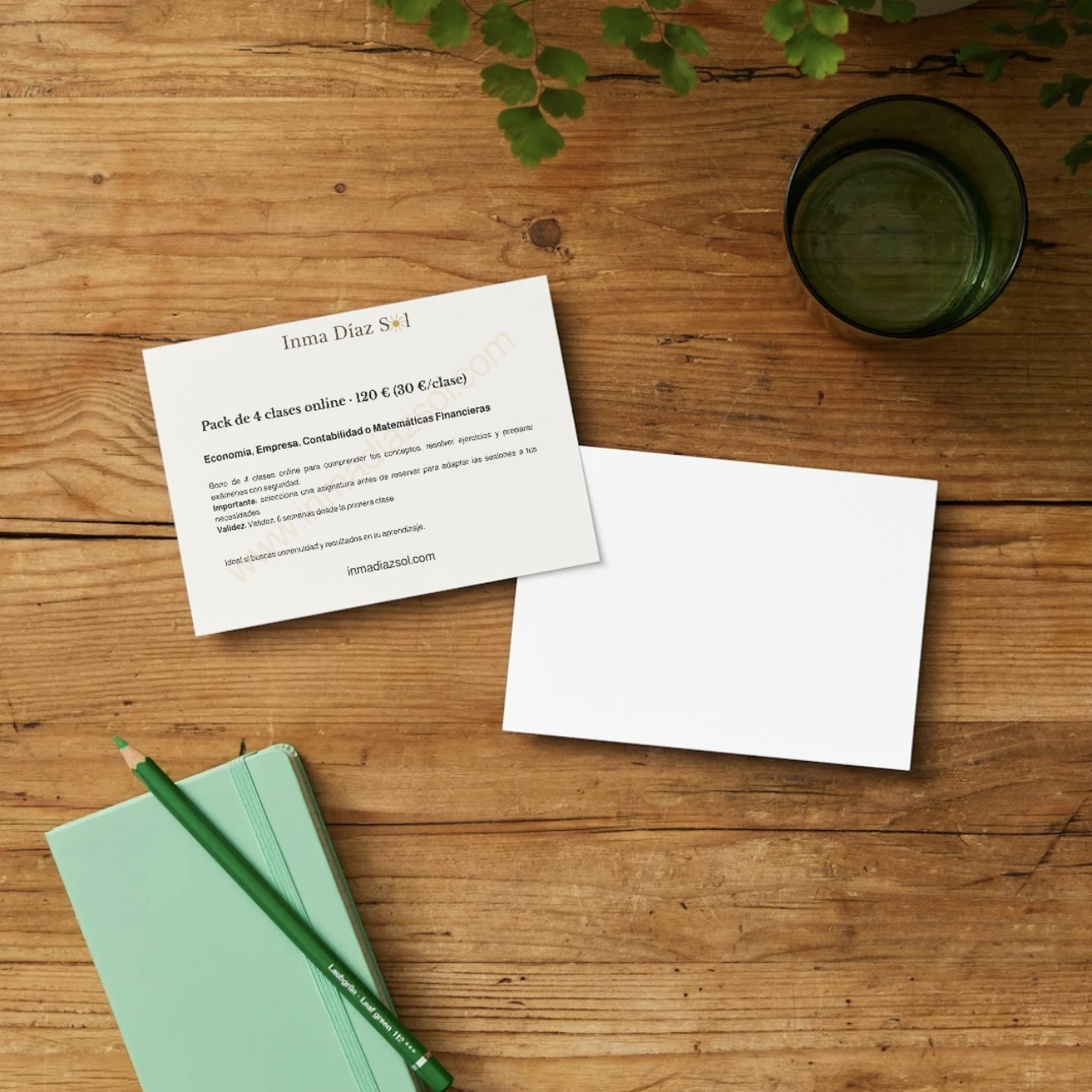 Material de estudio en una mesa de madera, incluyendo un folleto de 'Inma Díaz Sol' sobre clases en línea de economía, empresa, contabilidad y matemáticas financieras, un bloc de notas verde con un bolígrafo a juego, una hoja en blanco y un vaso con líquido