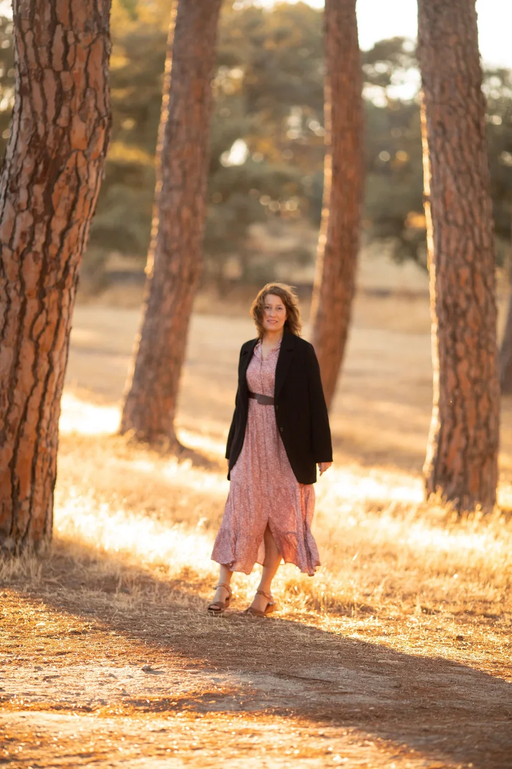 Mujer caminando por un bosque con árboles altos, vestida con vestido largo rosa y chaqueta negra, durante el atardecer