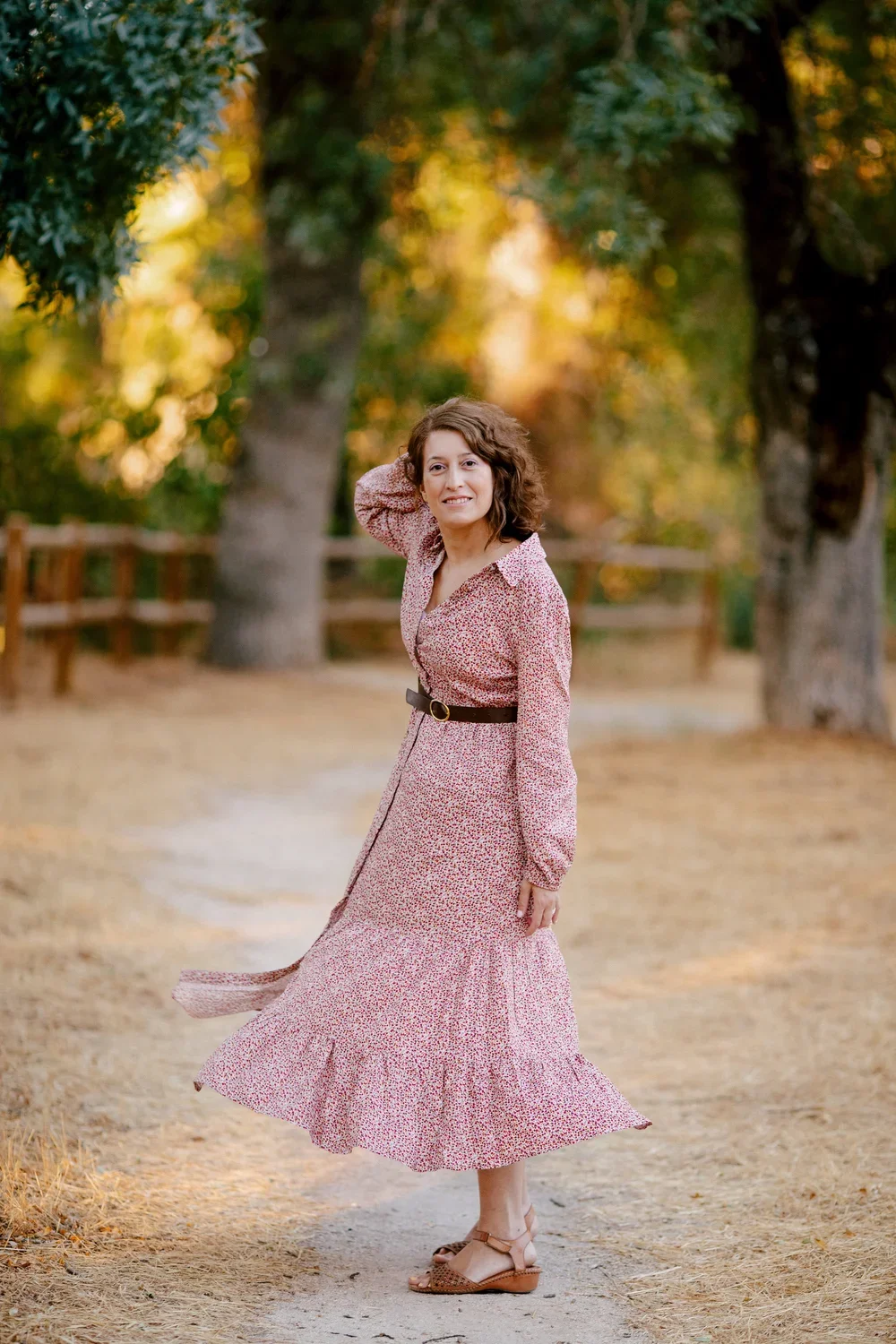 Una mujer con vestido largo de estampado floral y cinturón oscuro, caminando por un camino en un parque con árboles y hojas en tonos otoñales, durante la tarde.