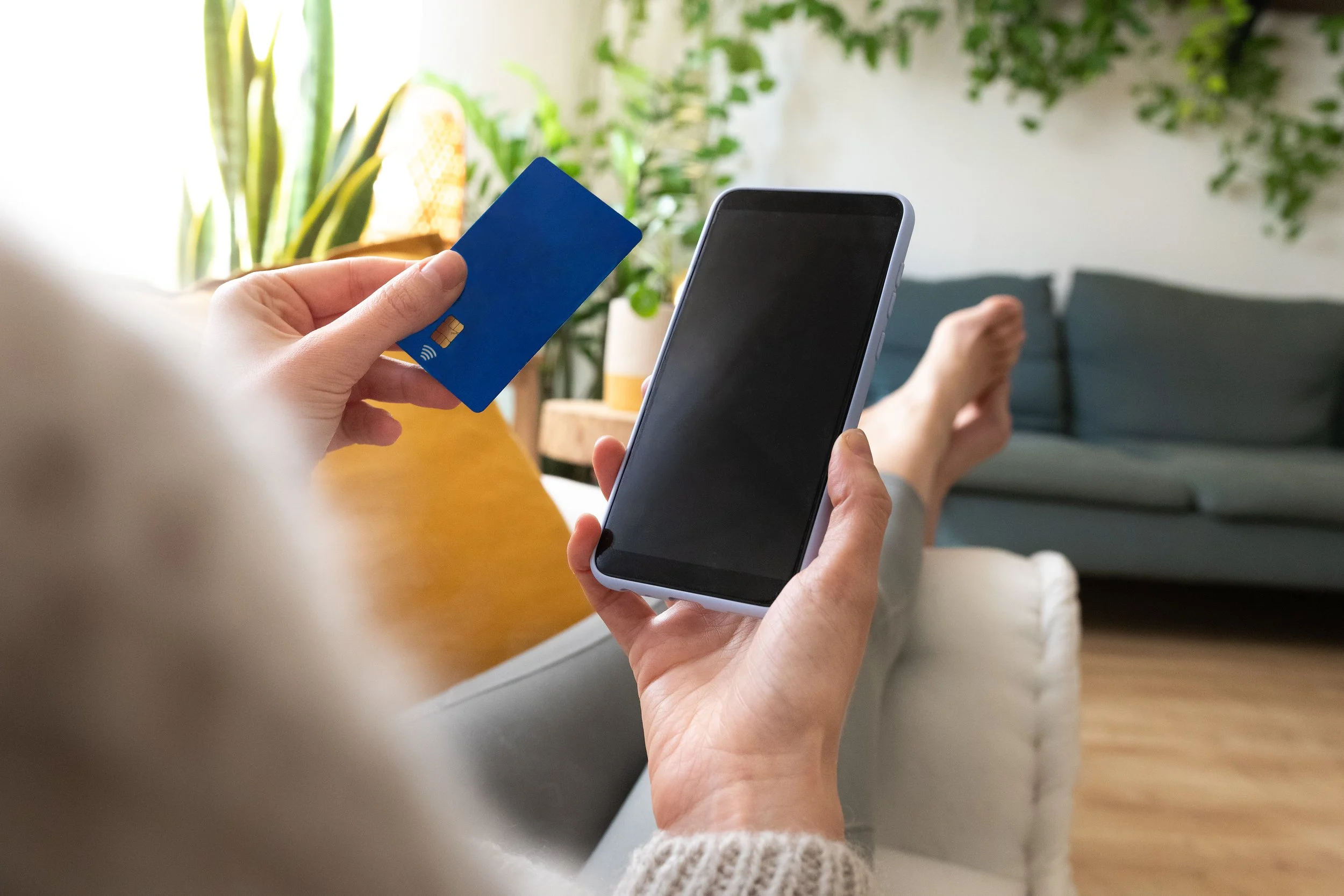 Persona usando un teléfono móvil y una tarjeta de crédito en una sala de estar con plantas y muebles.