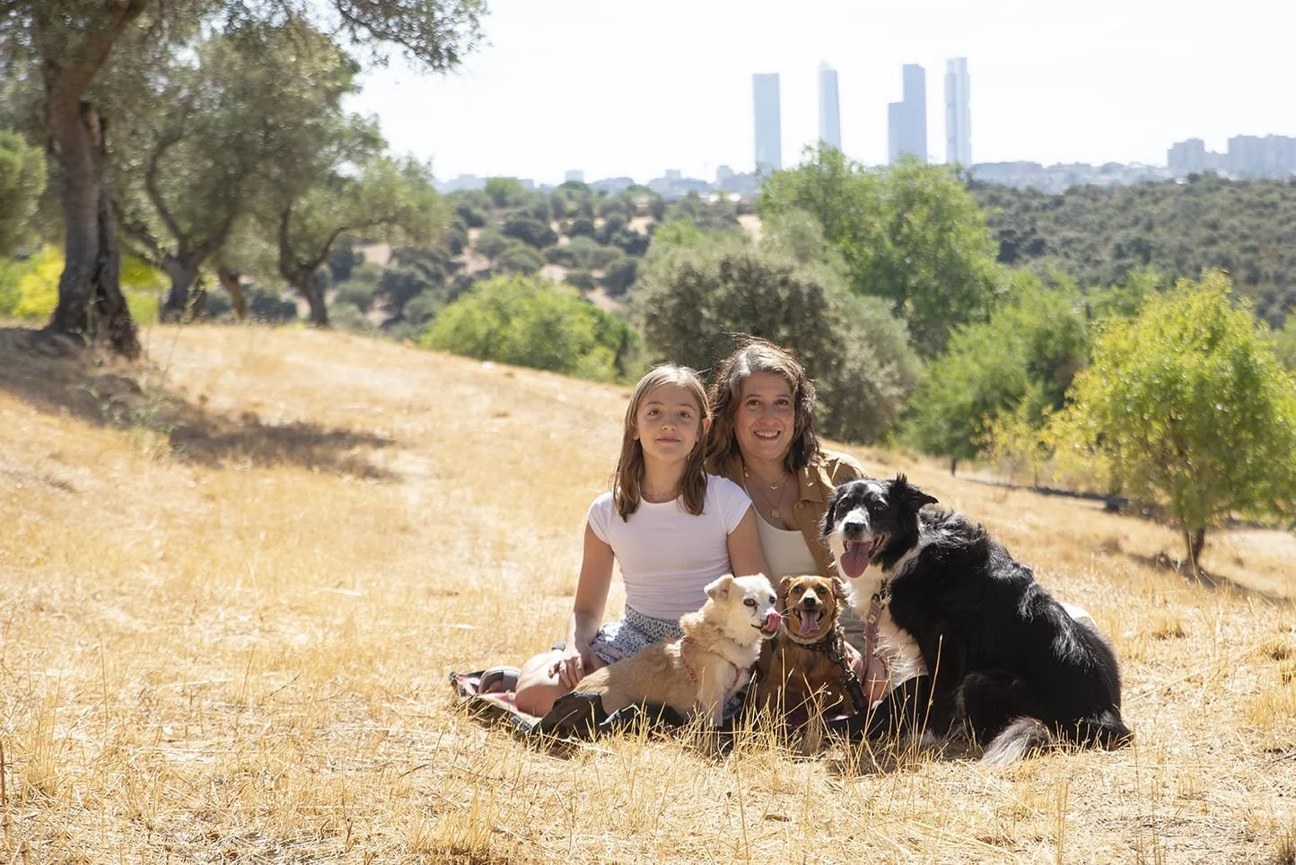 Mujer y niña con perros en un campo seco con árboles y ciudad al fondo.