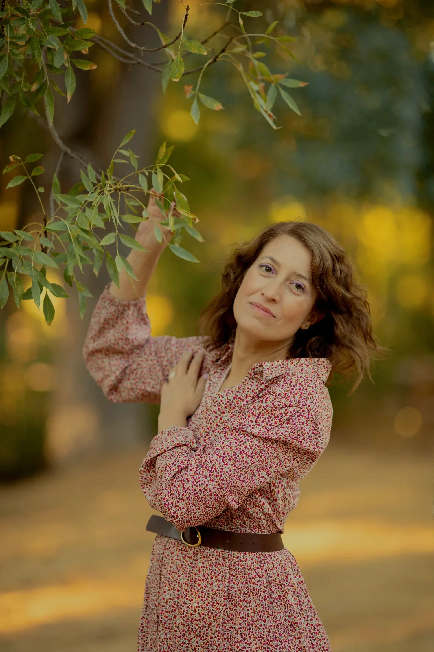 Mujer con cabello rizado, vistiendo un vestido con estampado de pequeñas flores, posando en un parque otoñal, tocando una rama de árbol.