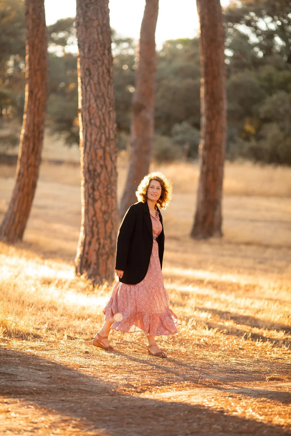 Mujer caminando por un bosque en la tarde, con árboles altos y sol brillante.
