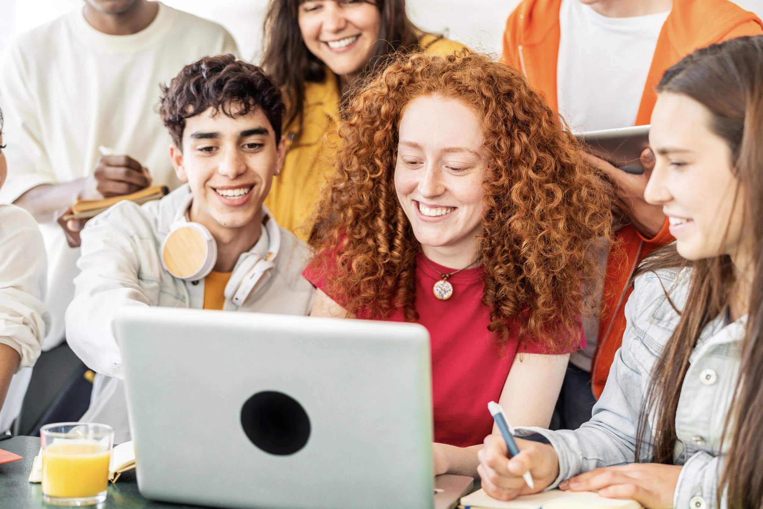Grupo de jóvenes y una mujer rubia con cabellera rizada, todos sonriendo y viendo una computadora portátil en una mesa, en un entorno de estudio o trabajo en equipo.