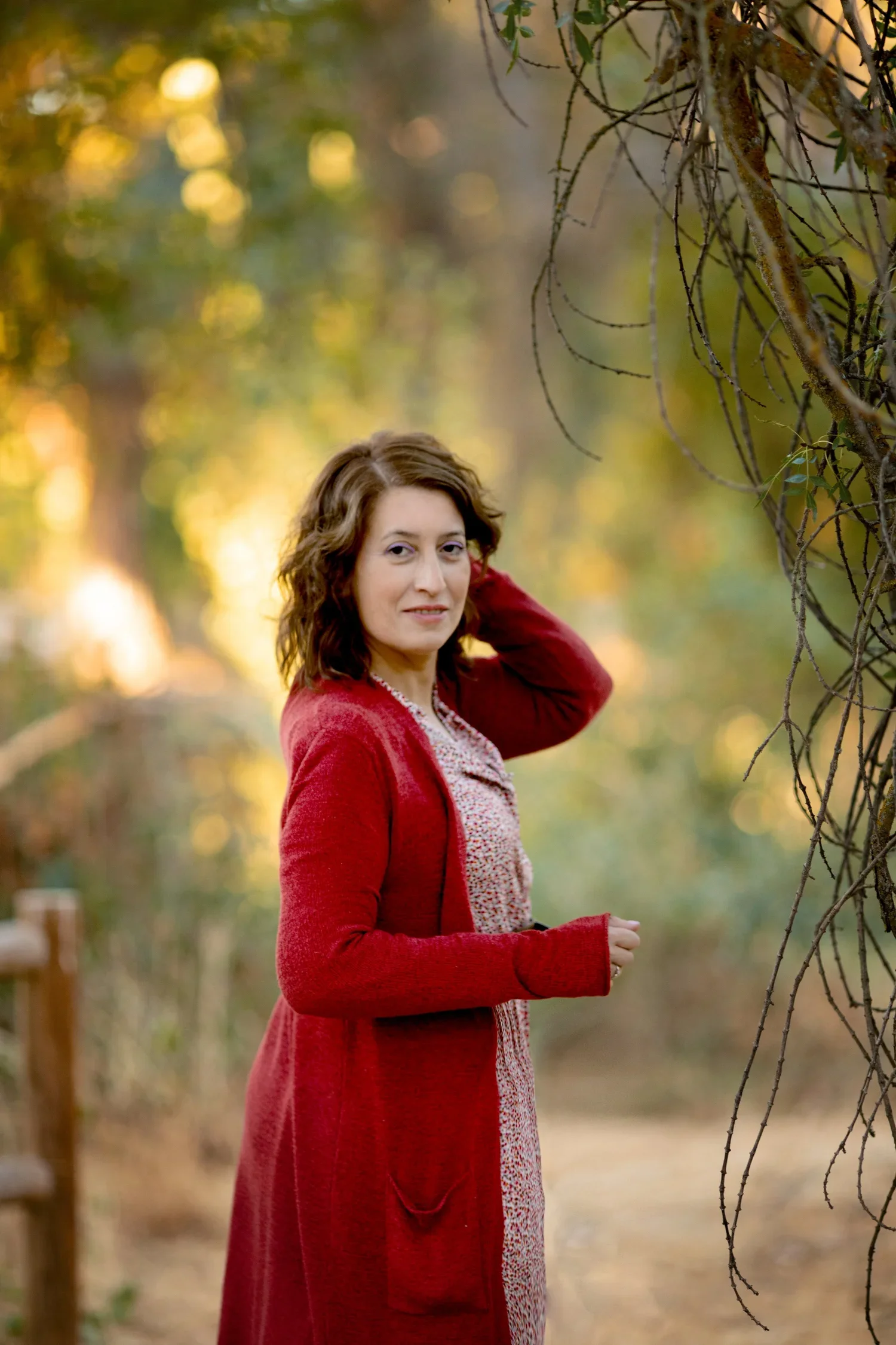 Mujer con cabello rizado, vestida con un abrigo rojo, en un entorno natural con árboles y ramas en el fondo durante el atardecer.