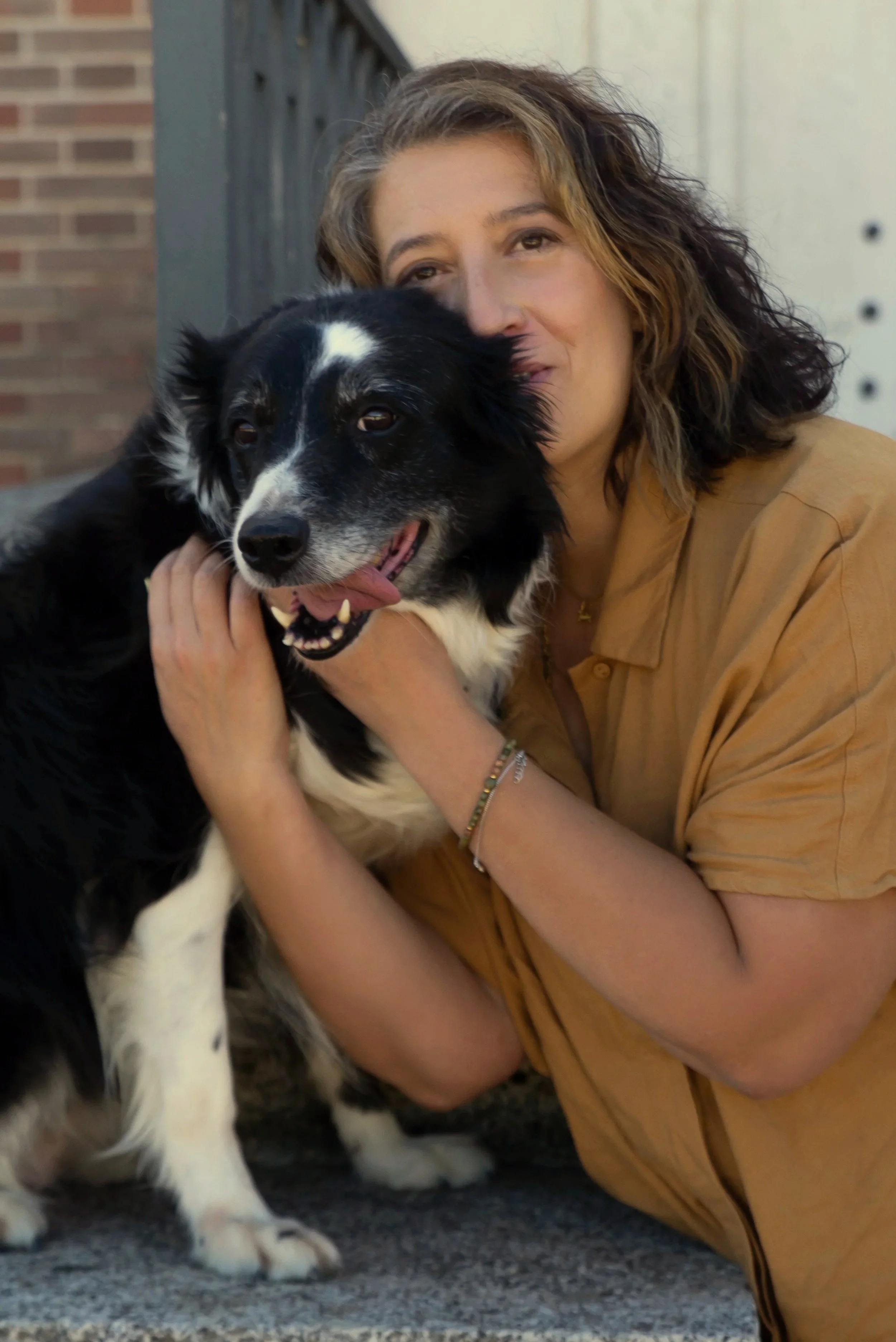 Una mujer con cabello rizado y de cabello castaño claro abraza a un perro border collie negro y blanco en una calle urbana.