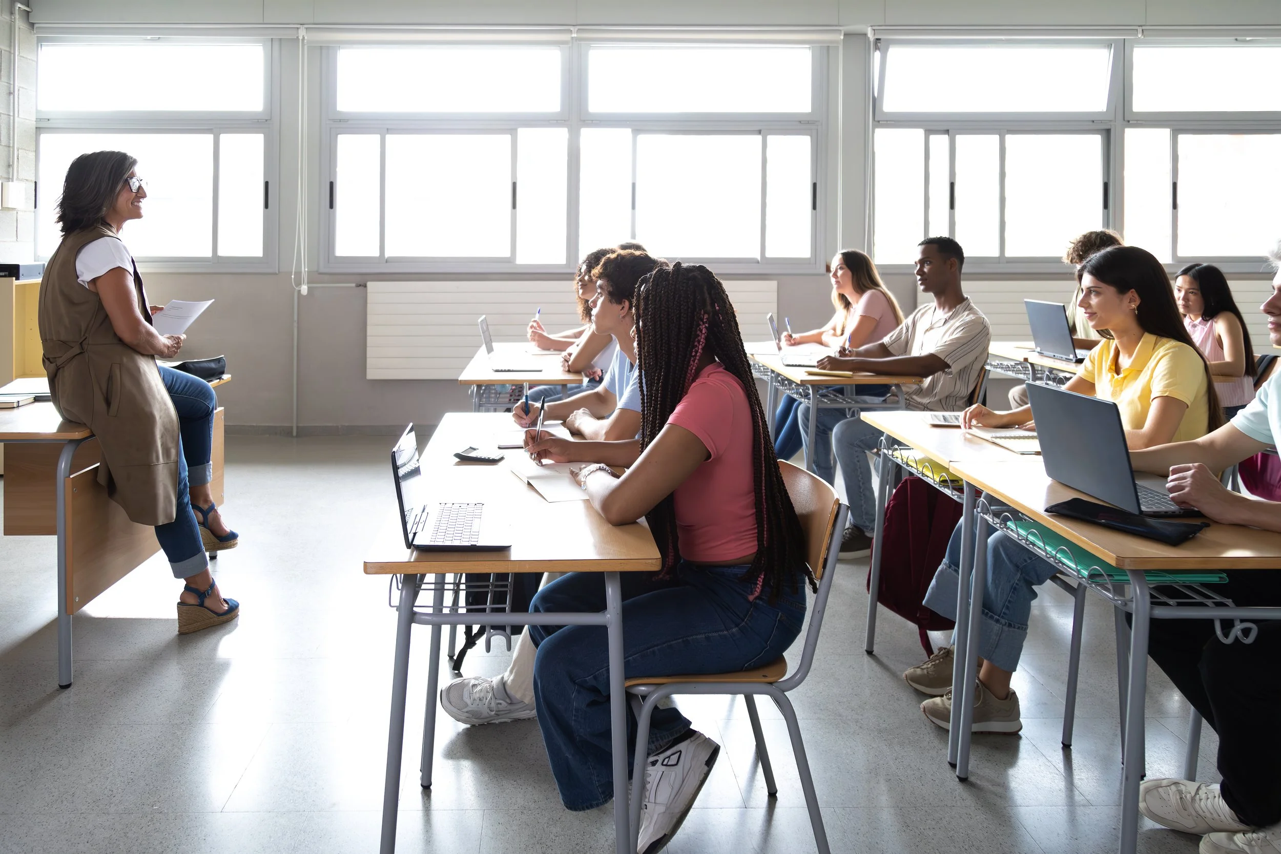 Clases en un salón con estudiantes sentados en escritorios con laptops, atendiendo a una profesora que está de pie frente a ellos, en una sala luminosa con ventanas grandes.