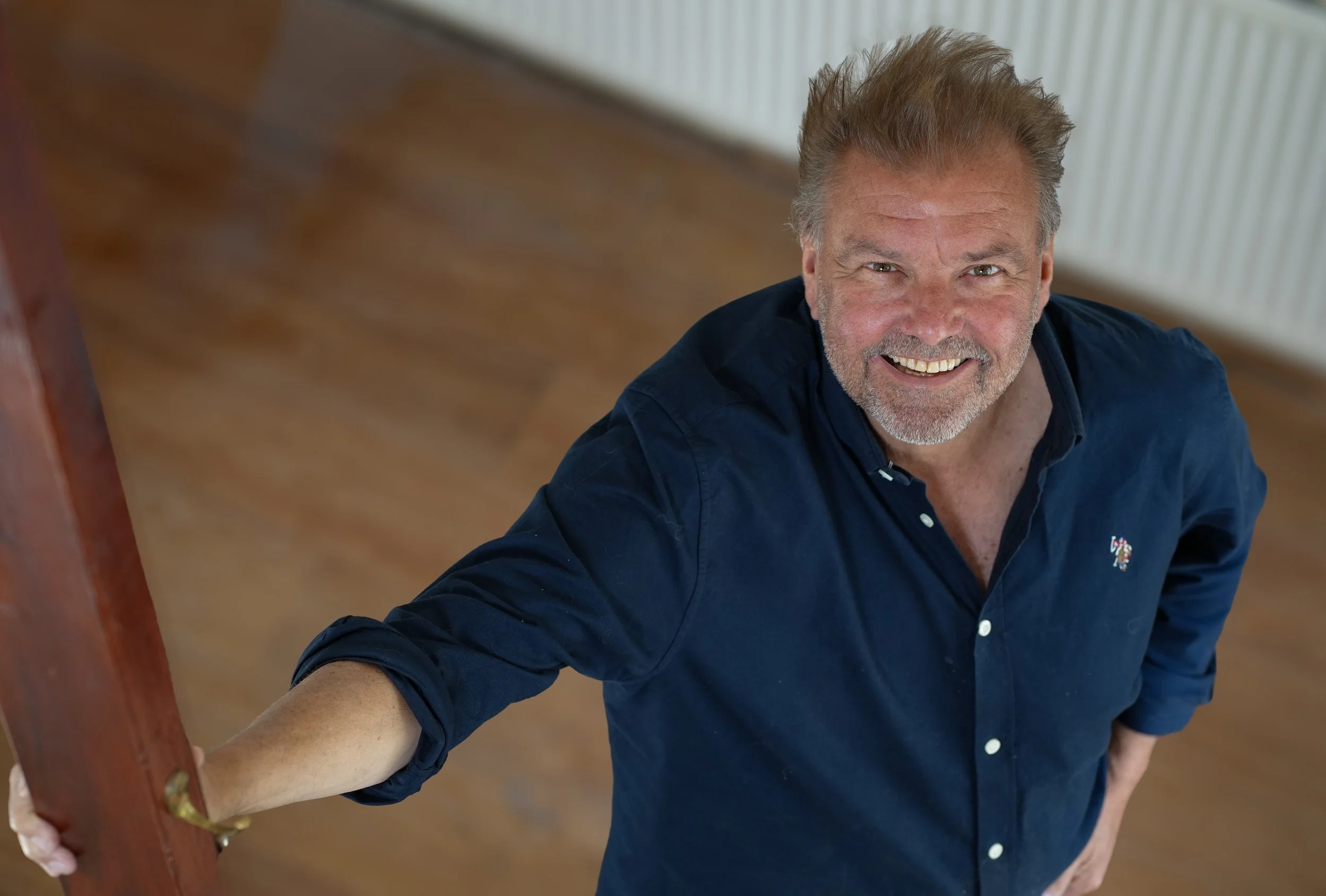 A smiling middle-aged man with gray hair and beard, wearing a navy blue shirt, is looking up at the camera and standing on a wooden floor, holding onto a wooden railing.
