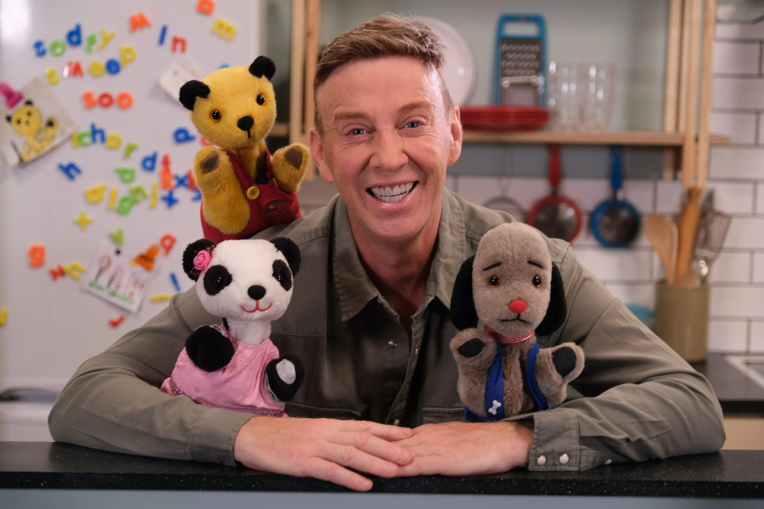 A smiling man sits at a kitchen counter with four stuffed animal puppets on his arms and shoulders, including a yellow bear, a panda, a gray dog, and a brown dog with a red nose.