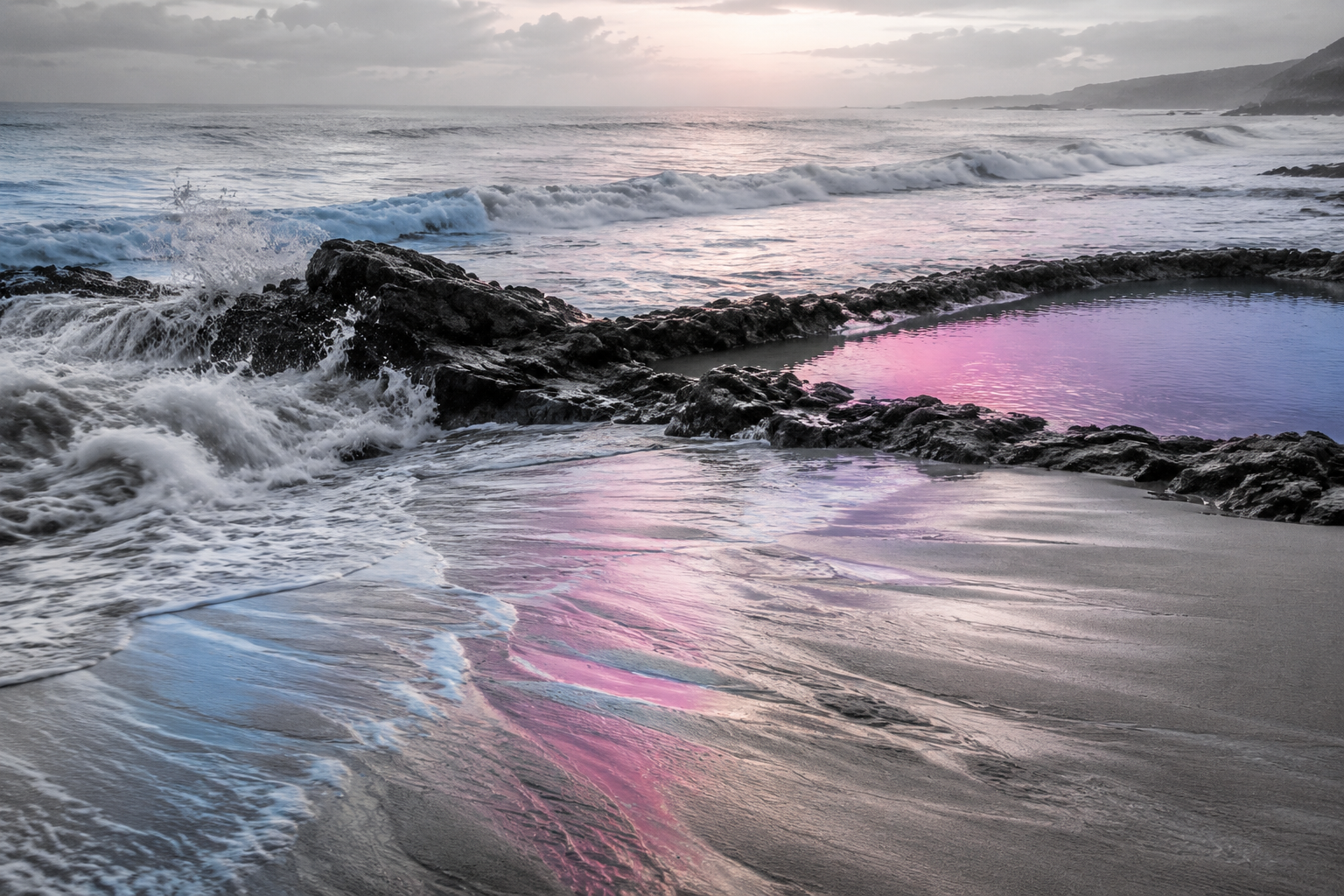 Ocean waves move around dark coastal rocks as soft blue and pink light reflects across a quiet tidal pool and wet sand.