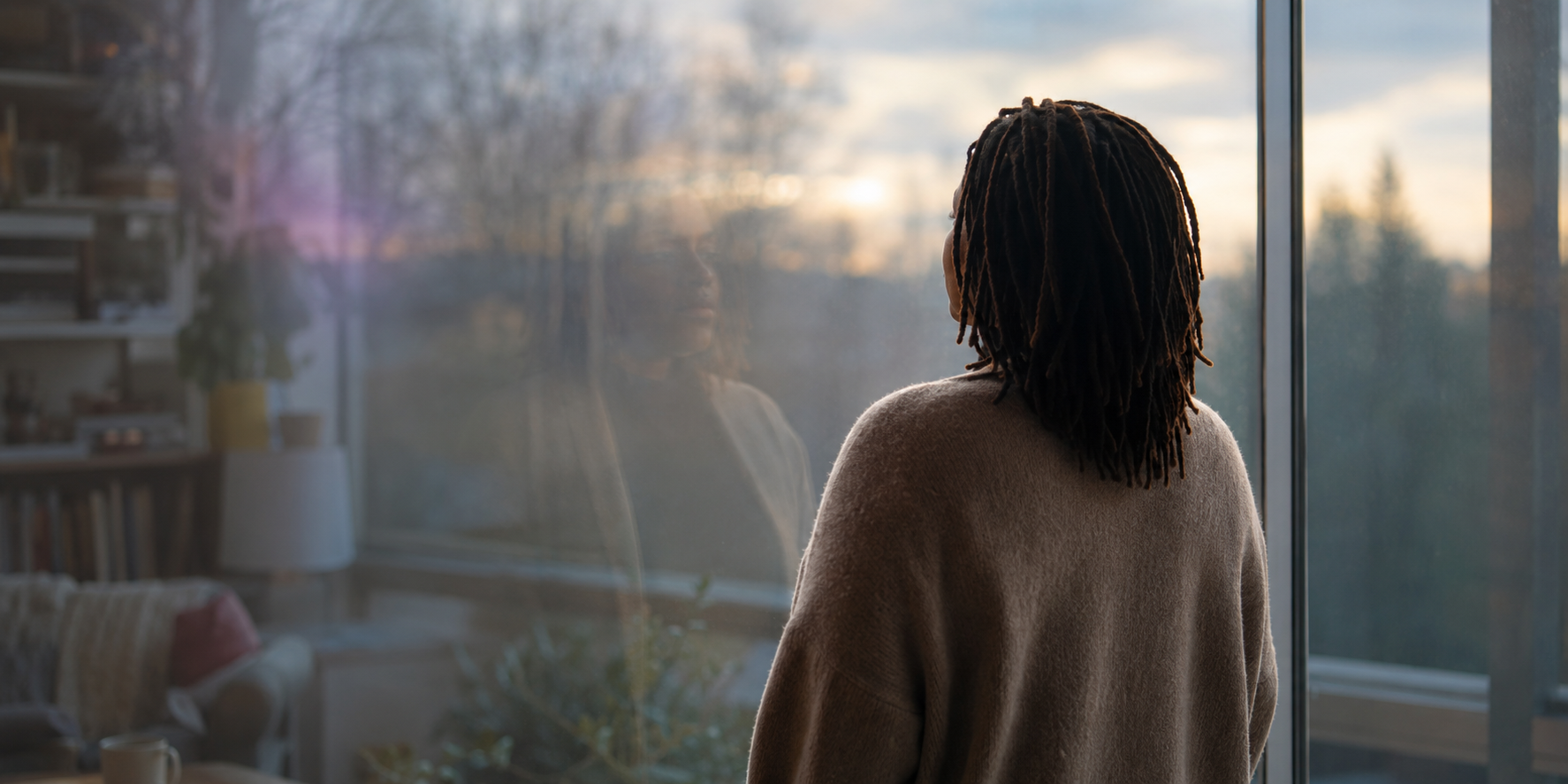 An Black woman with shoulder-length locs stands before a large window, looking toward a softly lit horizon. Her reflection and the room behind her appear faintly in the glass.