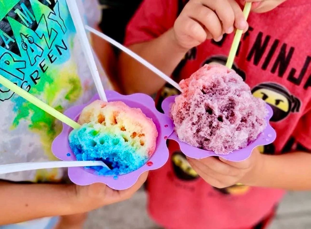 Child holding two purple silicone cupcake liners filled with shaved ice, one colorful rainbow and one pink, each with a straw for drinking.
