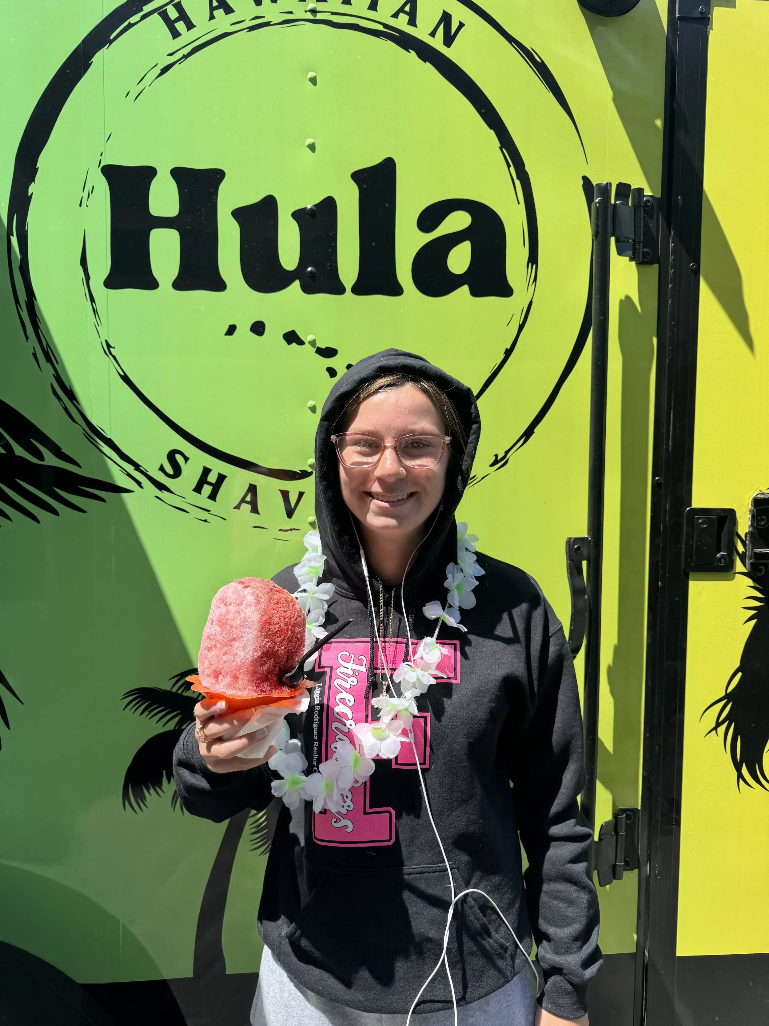 A young woman wearing glasses, a black hoodie with pink lettering, and a Hawaiian lei, holding a shaved ice cone with a pink topping, standing in front of a bright green food truck with the word 'Hula' on it.