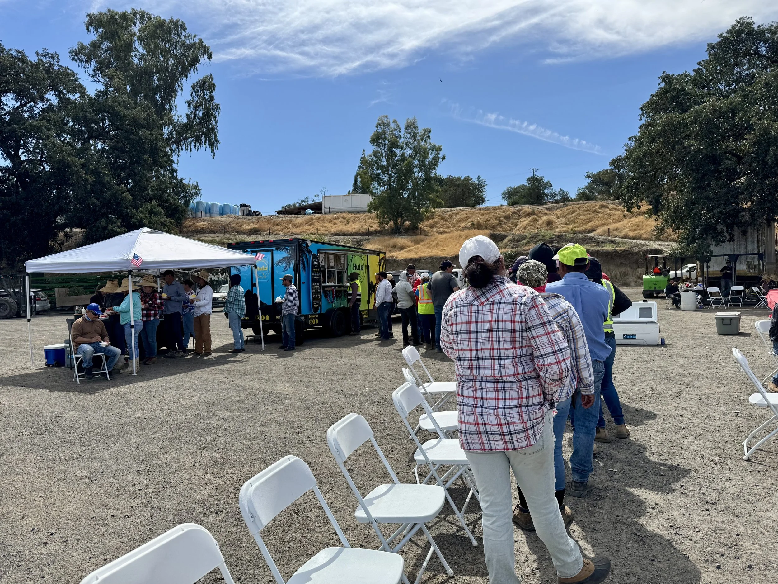 Line of people waiting outdoors near a food truck on a sunny day. Some chairs are set up nearby, with trees and a hillside in the background.