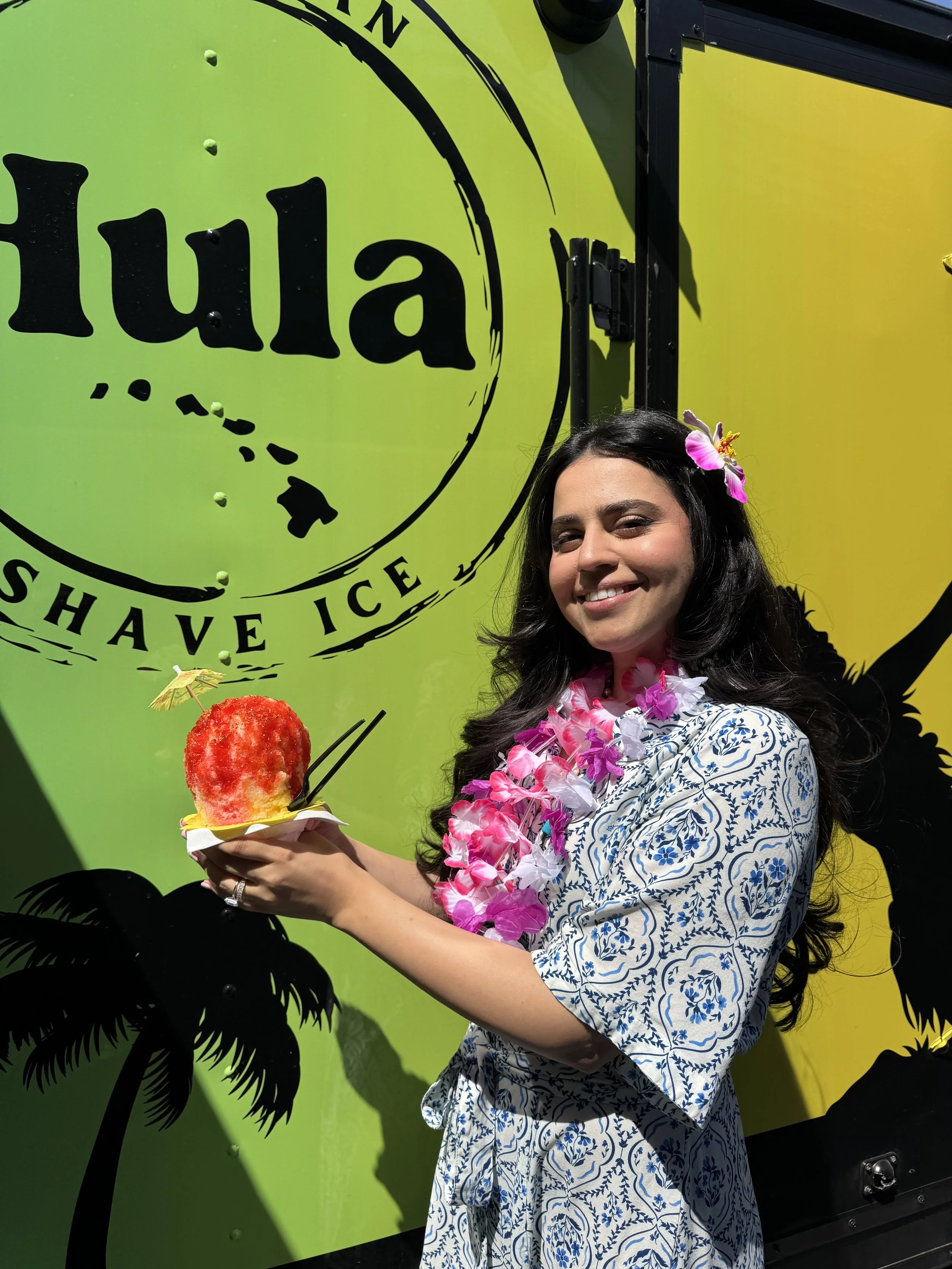 A woman with dark hair wearing a floral lei and dress, holding a shaved ice dessert with a umbrella and tropical decorations, standing in front of a green and yellow wall with the logo 'Hula' and a palm tree silhouette.