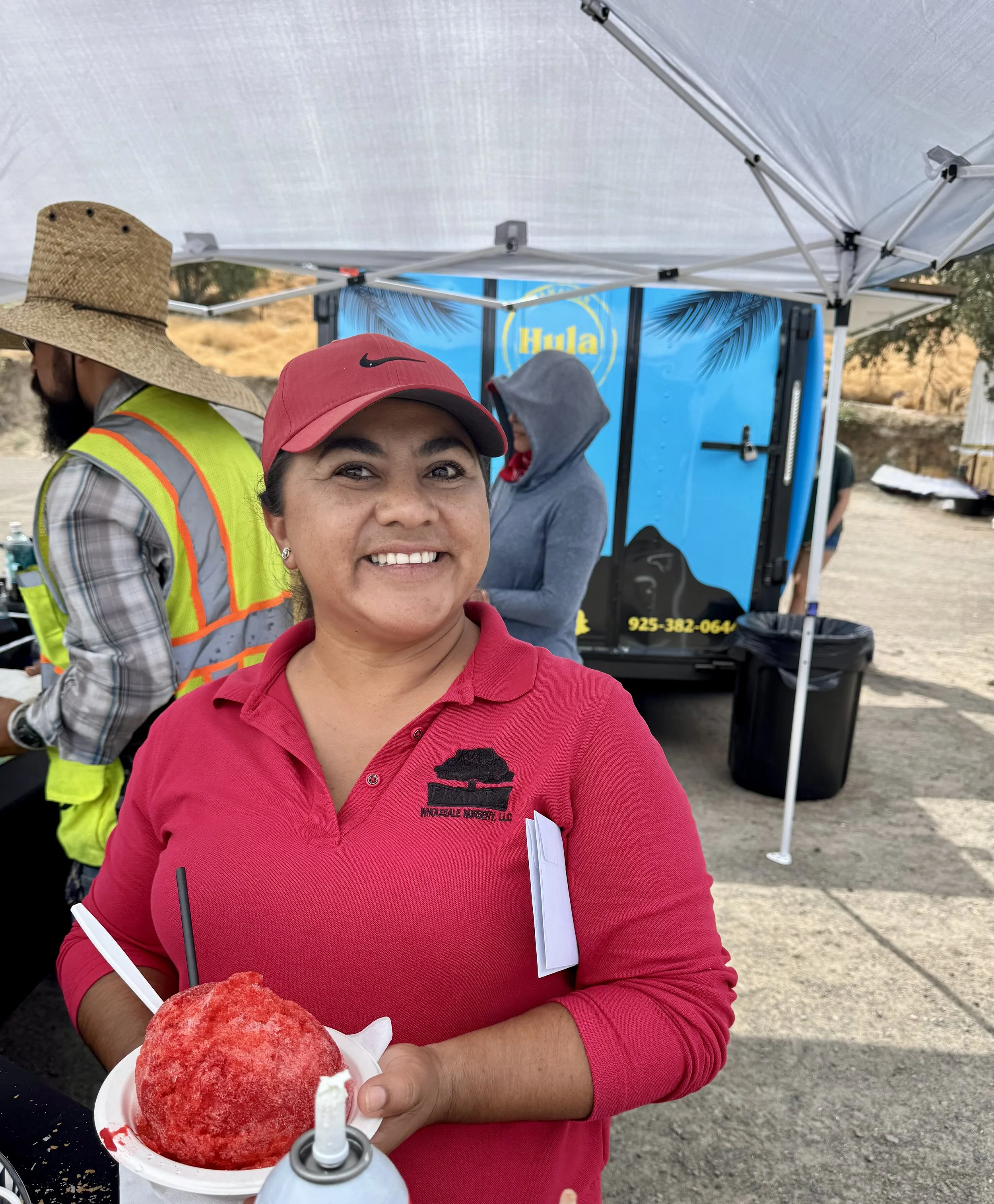 Woman smiling holding a cup of shaved ice with red syrup at an outdoor event, with a carnival or food truck setup in the background.