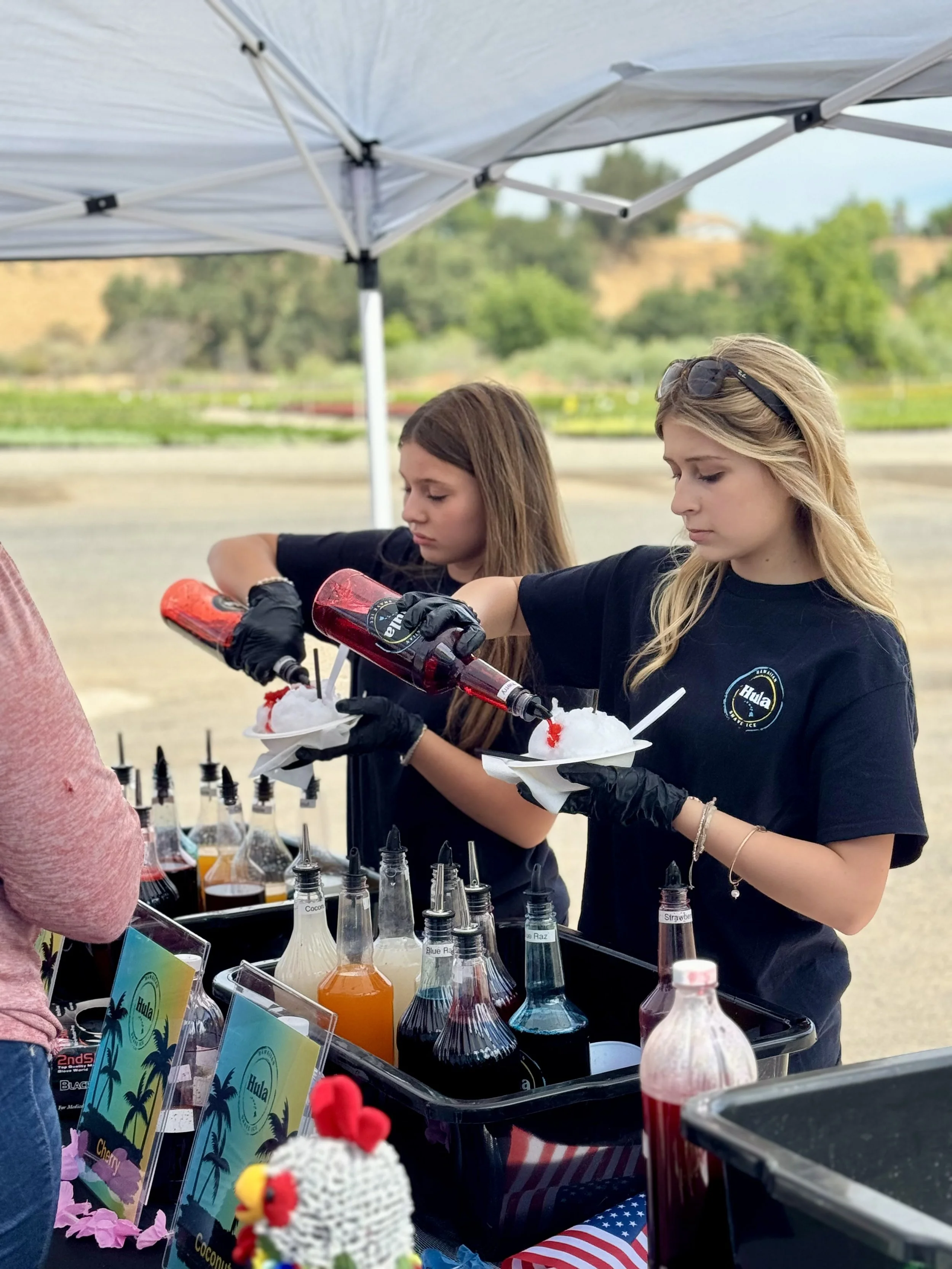 Two young women in black shirts preparing snow cones with flavored syrup at an outdoor stand under a canopy, with colorful syrup bottles and signs, on a bright day with green scenery in the background.