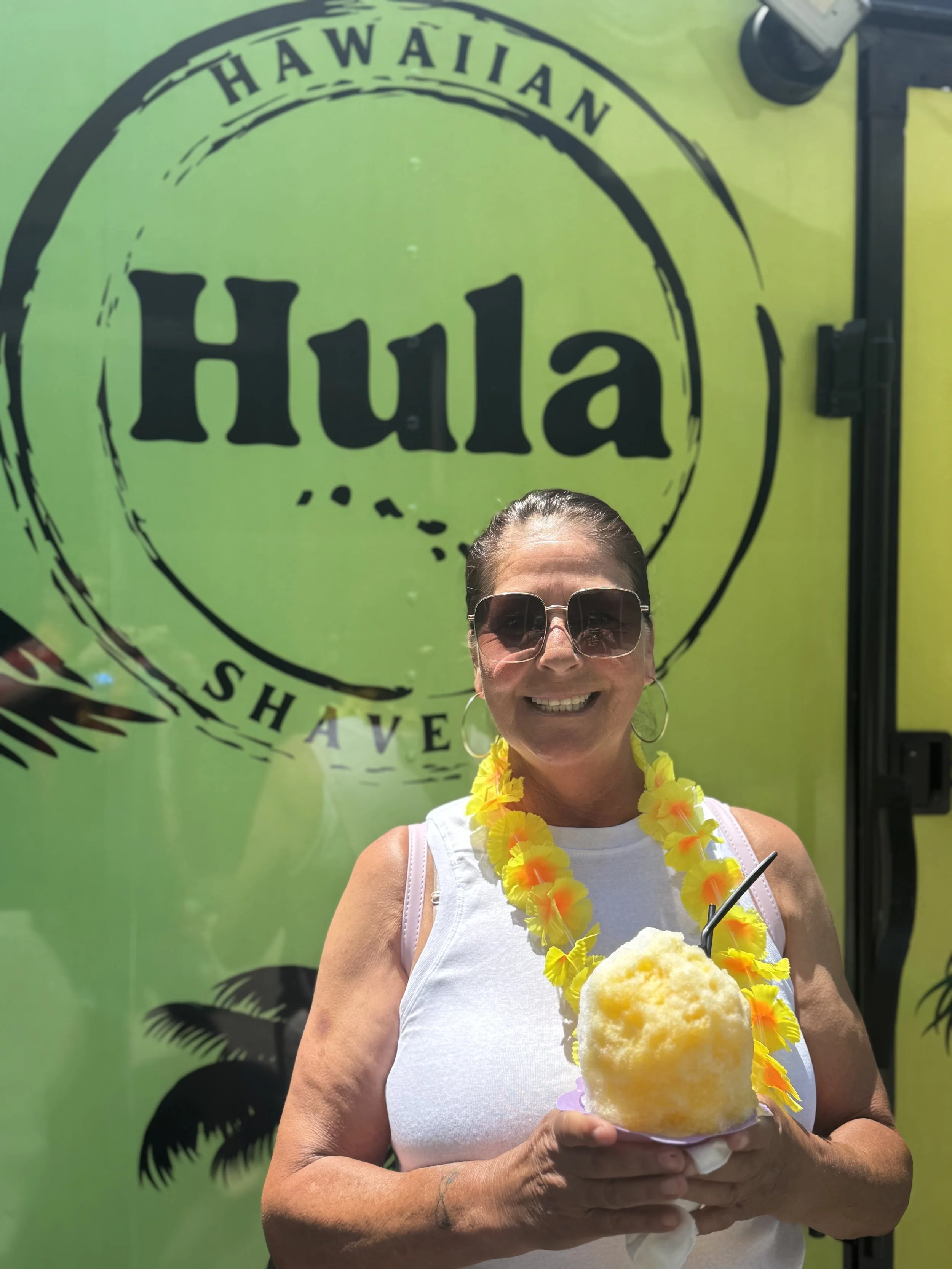 A woman smiling while holding a yellow snow cone in front of a green sign that says 'Hawaii Shave'. She is wearing sunglasses, a white sleeveless top, a yellow flower lei, and hoop earrings.