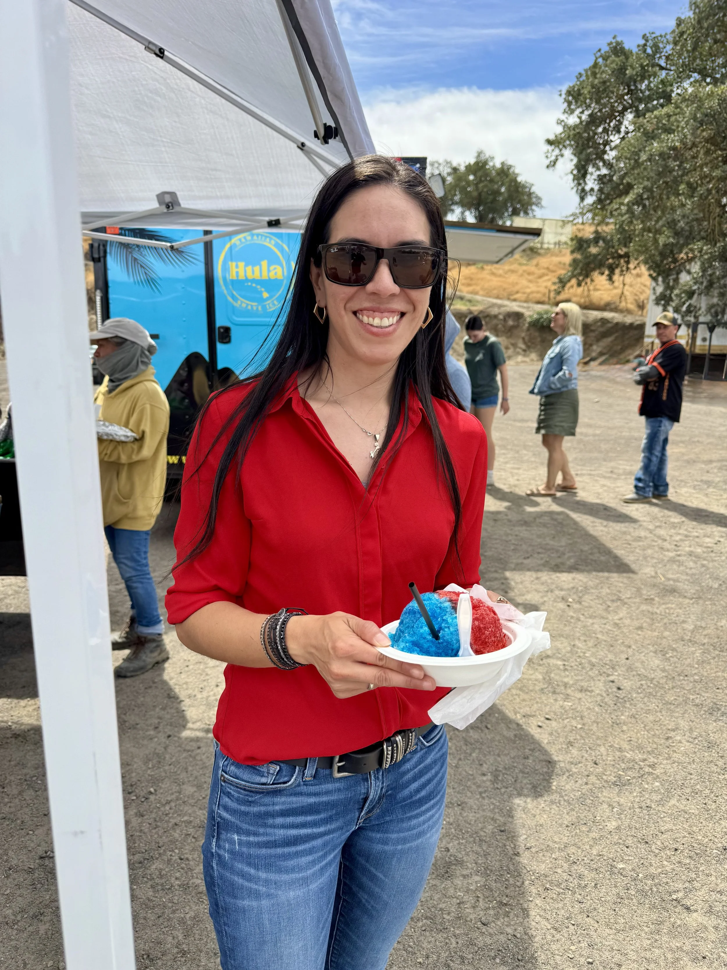 A woman wearing sunglasses and a red shirt holding a plate of blue and red shaved ice at an outdoor event on a sunny day.