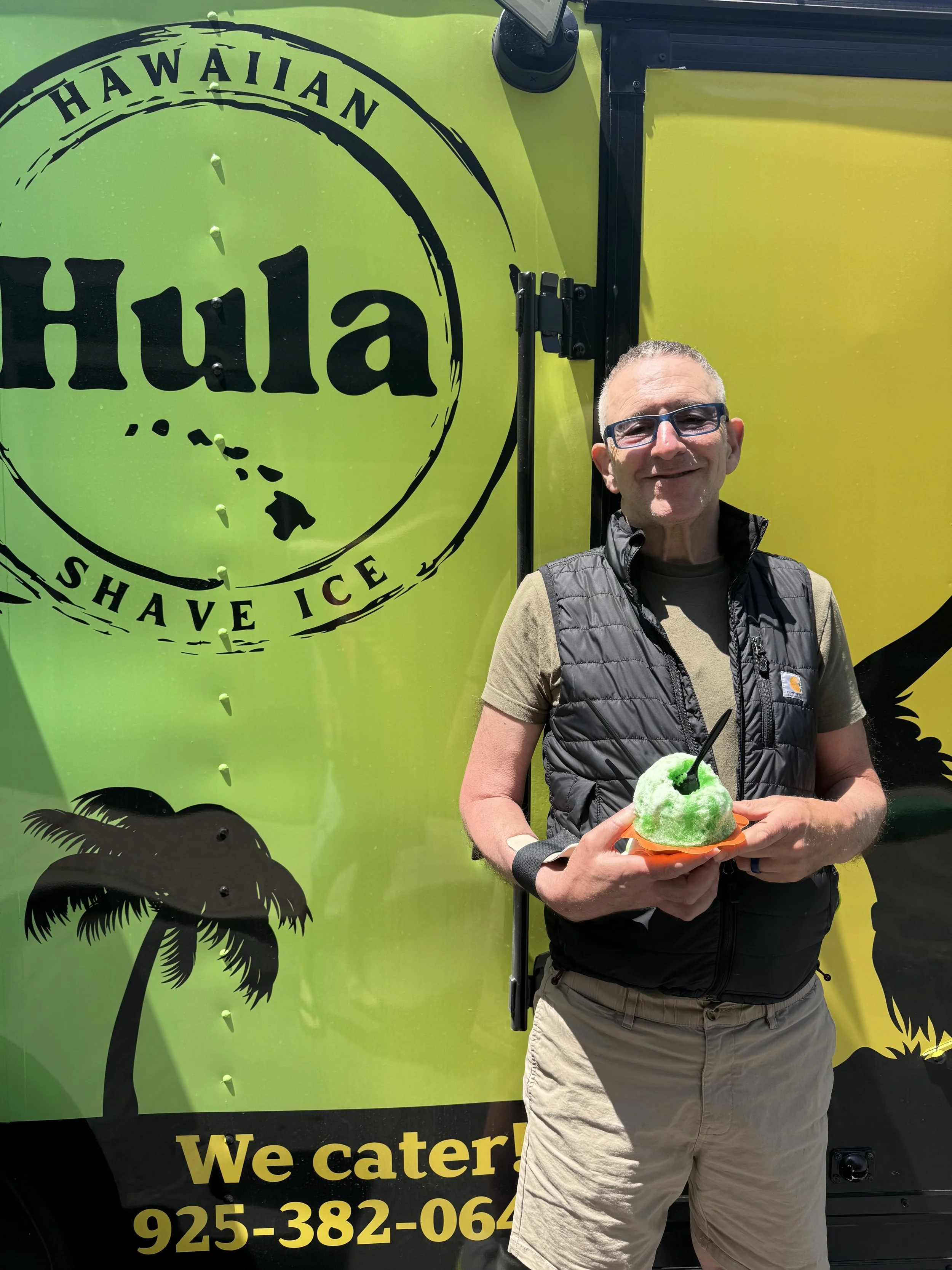 A man holding a green shaved ice dessert with a black straw and spoon, standing in front of a bright green Hawaiian shave ice truck with a palm tree graphic and the words "Hawaiian Shave Ice" and "We cater!"