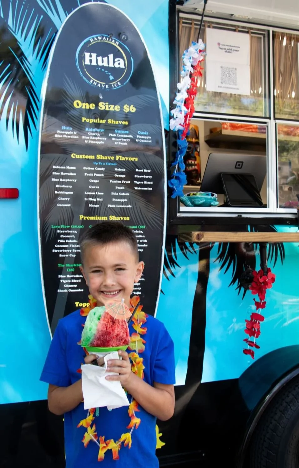 A young boy smiling and holding a colorful shaved ice dessert at a Hawaiian-themed food truck. The truck has a menu board with shaved ice flavors and toppings, decorated with leis and luau-themed decorations.