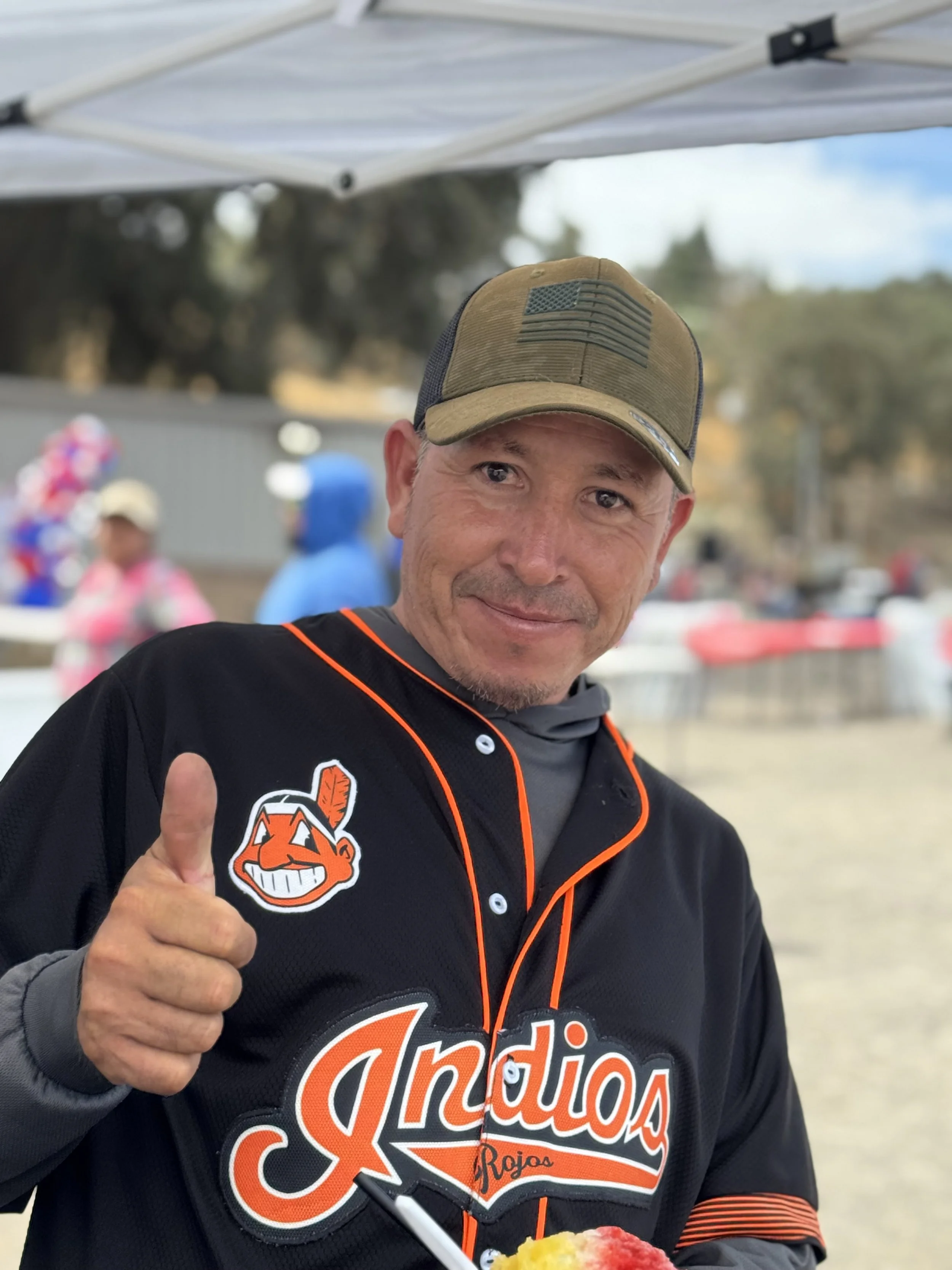 A man wearing a black Cleveland Indians baseball jersey and a tan cap, giving a thumbs-up at an outdoor event.