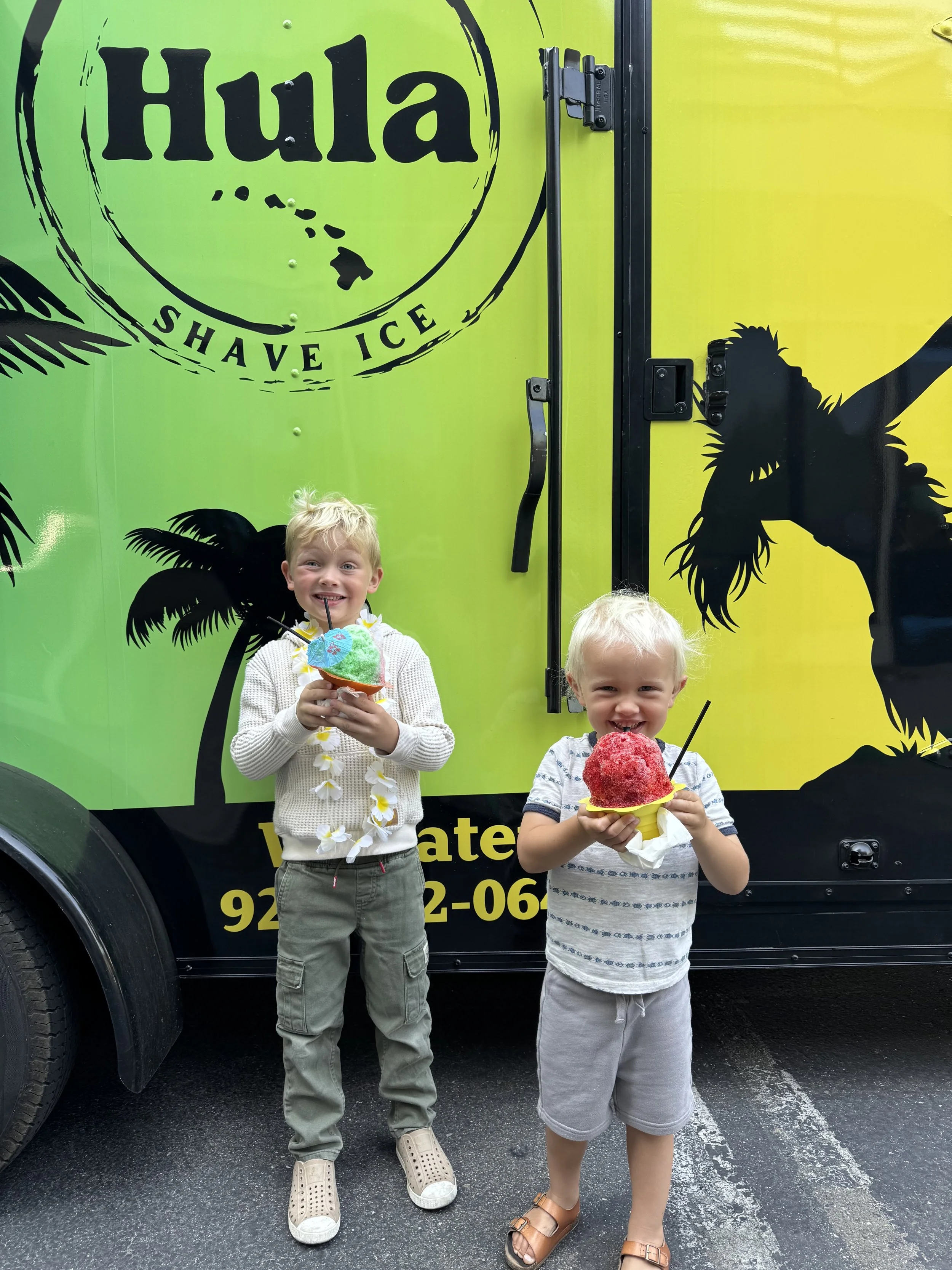 Two young boys standing in front of a green and black ice cream truck, each holding a colorful shaved ice treat and smiling.