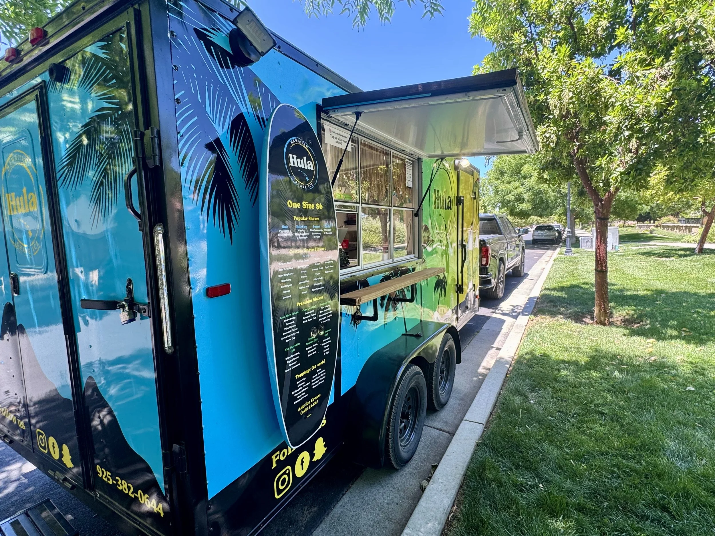 A colorful food truck parked on the side of a street with trees and grass, serving shaved ice, with menu displayed on the side.