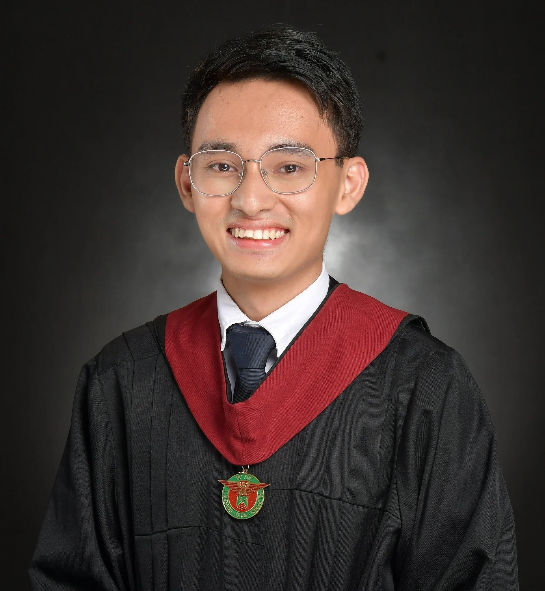Young man in a graduation cap and gown, wearing glasses, smiling, against a dark background.