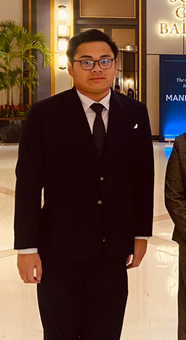 A young man wearing a black suit, white shirt, and patterned black tie standing indoors at a formal event.