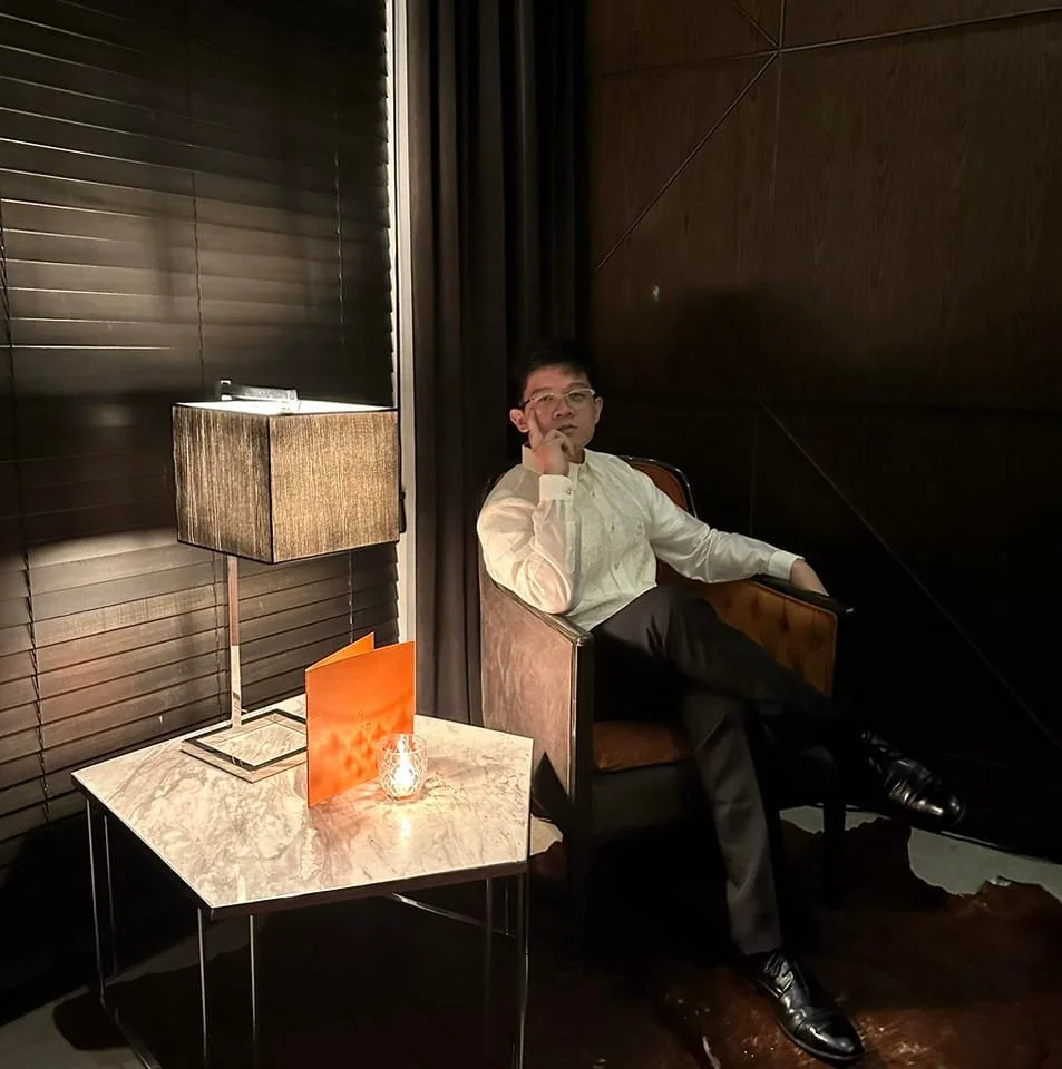 A man in a white shirt and black pants sitting in a brown armchair in a dimly lit room, with a side table with a lamp and a small candle holder, and dark wood walls.