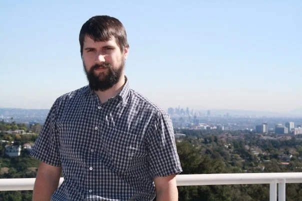 Young man with a beard standing on a balcony overlooking a cityscape, wearing a checked short-sleeve shirt.