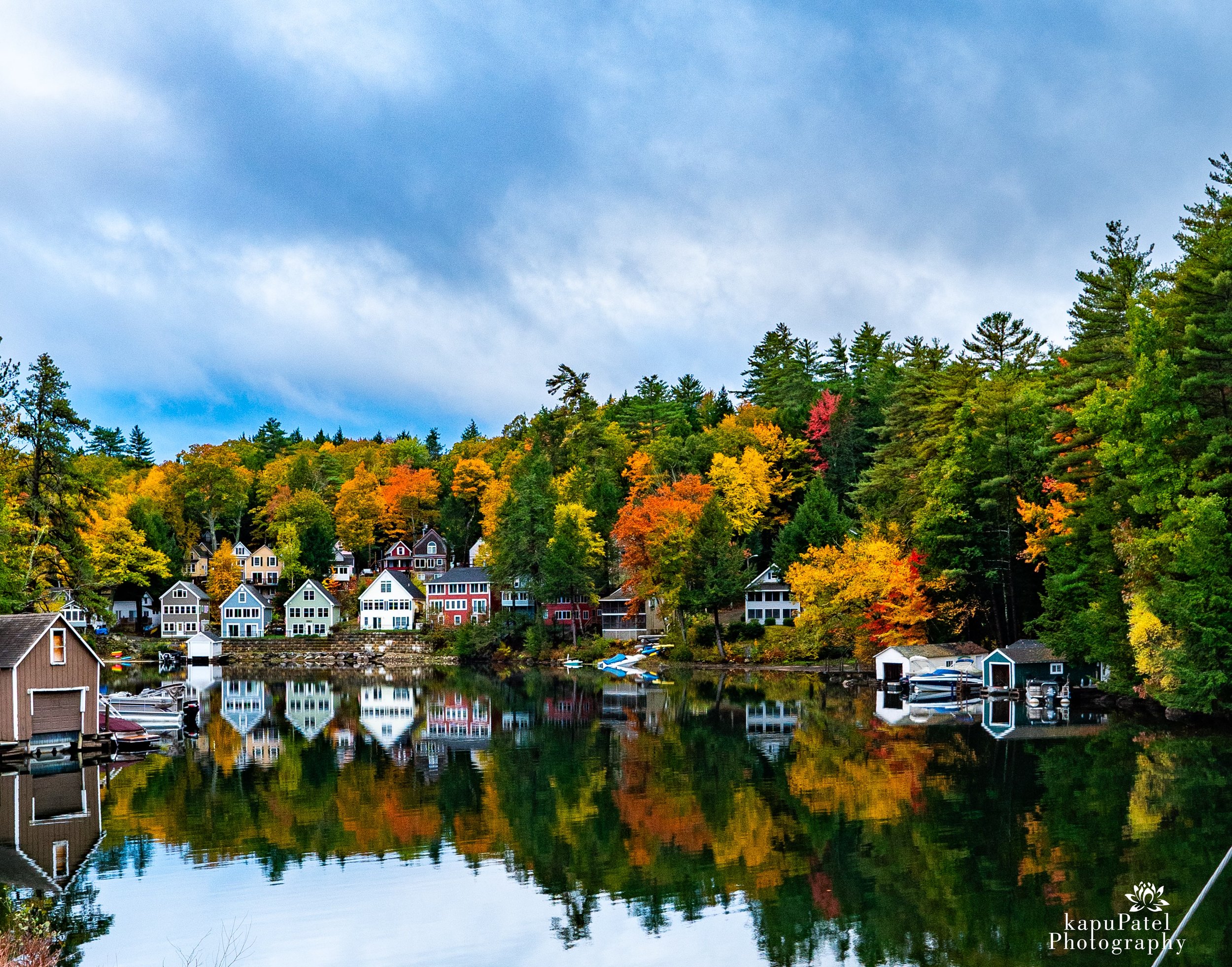 Colorful houses along a lake with trees displaying fall foliage, reflected in the water, under a partly cloudy sky.