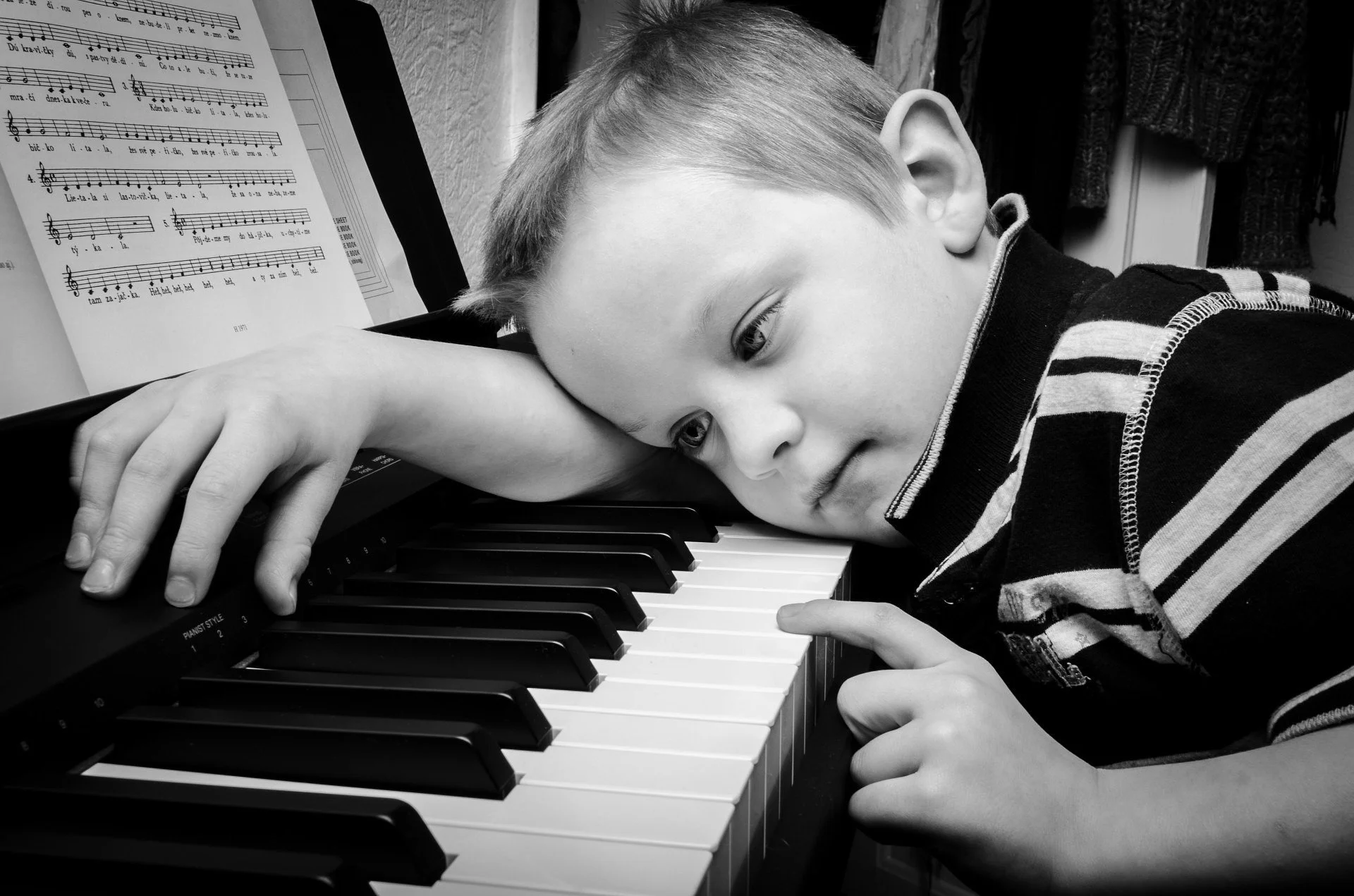 Un jeune garçon repose sa tête sur un clavier de piano, regardant le piano, avec une partition musicale devant lui.