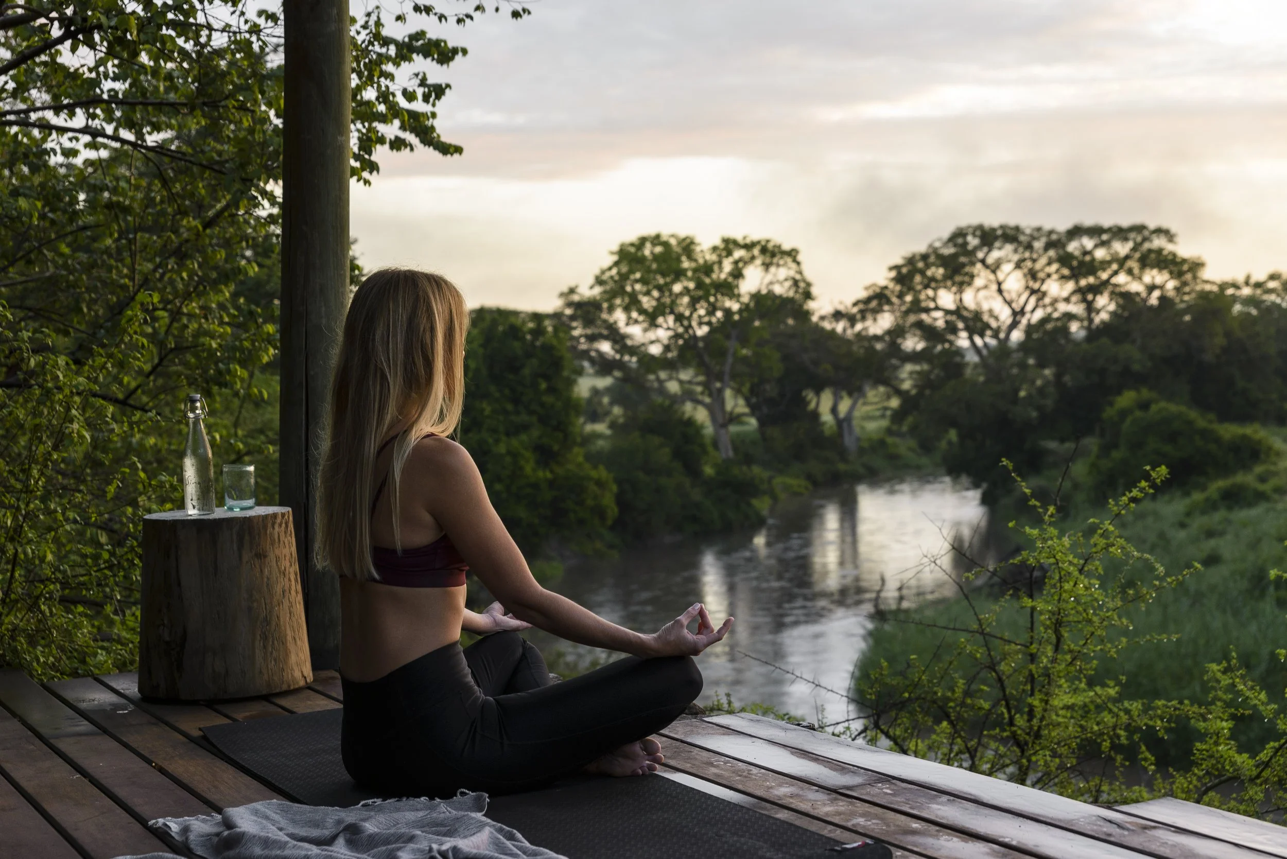 Eine Frau sitzt in einer Meditationshaltung auf einer wooden Plattform, blickt auf einen Fluss und umgeben von Bäumen, bei Sonnenuntergang in Afrika