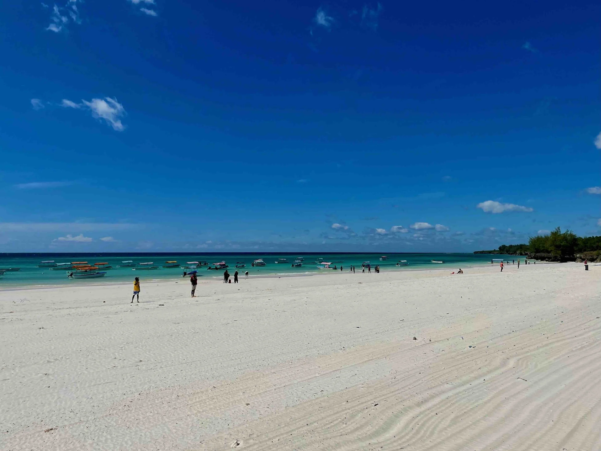 Weiße Strandküste mit mehreren Menschen, Boote im Wasser und einem klar blauen Himmel auf Sansibar