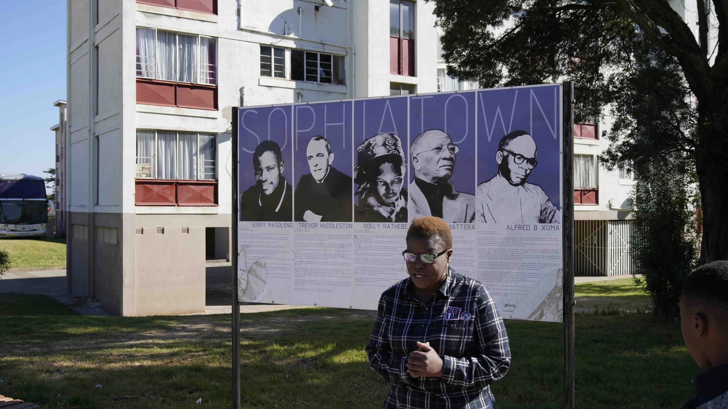 Eine Frau steht vor einem schwarzen Informationsschild mit Porträts von fünf bekannten Persönlichkeiten auf einem Rasen vor einem Wohnblock. Das Schild trägt den Titel "SOPHIALOW" in Johannesburg in Südafrika