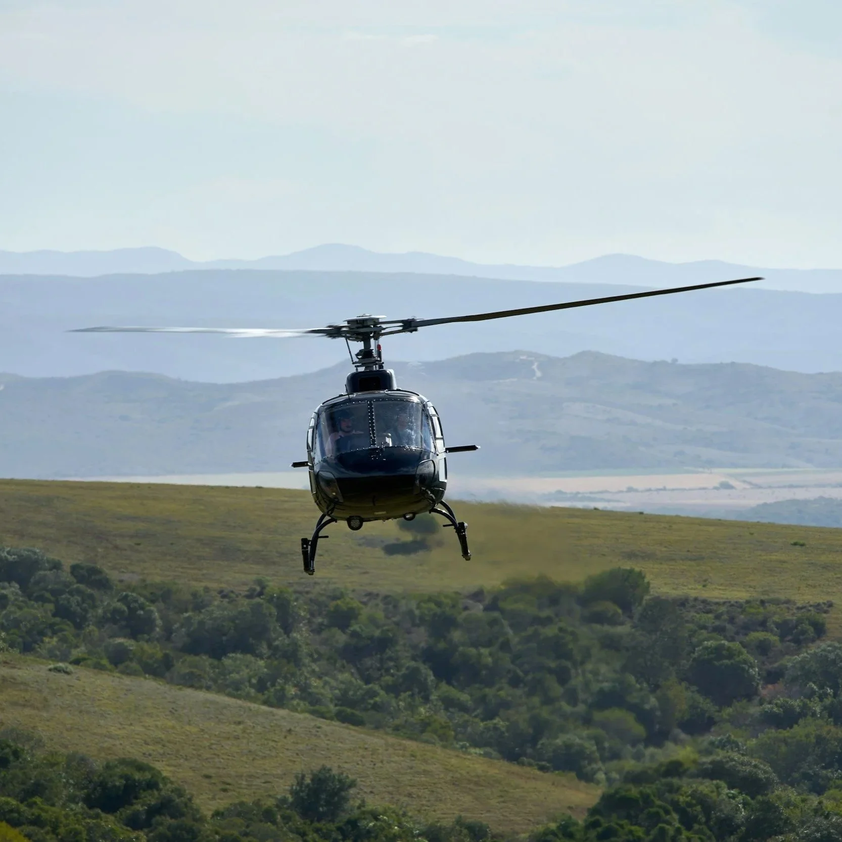 Ein schwarzer Hubschrauber fliegt über grüne Hügel mit Bäumen und Feldern, im Hintergrund Berge und Himmel in Afrika