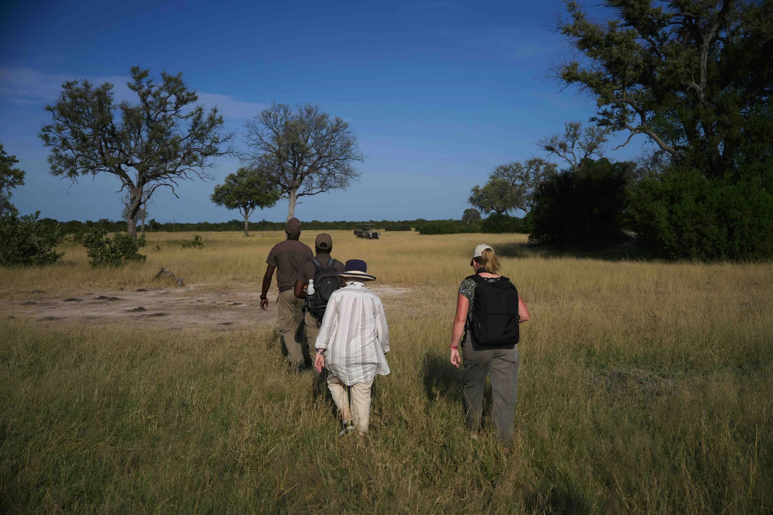 Menschen auf einer Walking Safari in Sambia in einer Steppenlandschaft. Im Hintergrund steht ein Safari Jeep. 