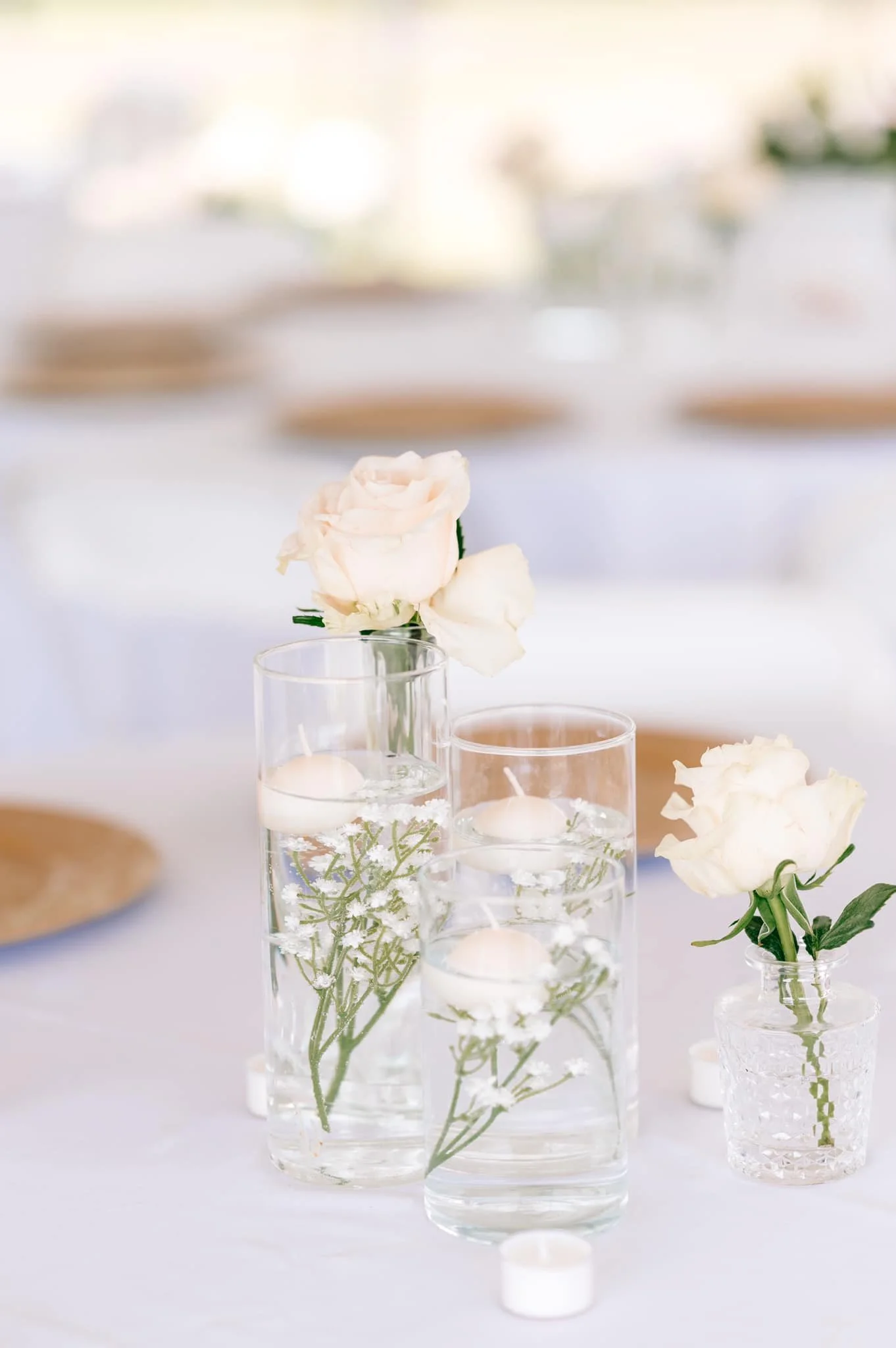 Elegant table centerpiece with three glass vases filled with water, pink and white roses, and floating candles, on a white tablecloth at a formal event.