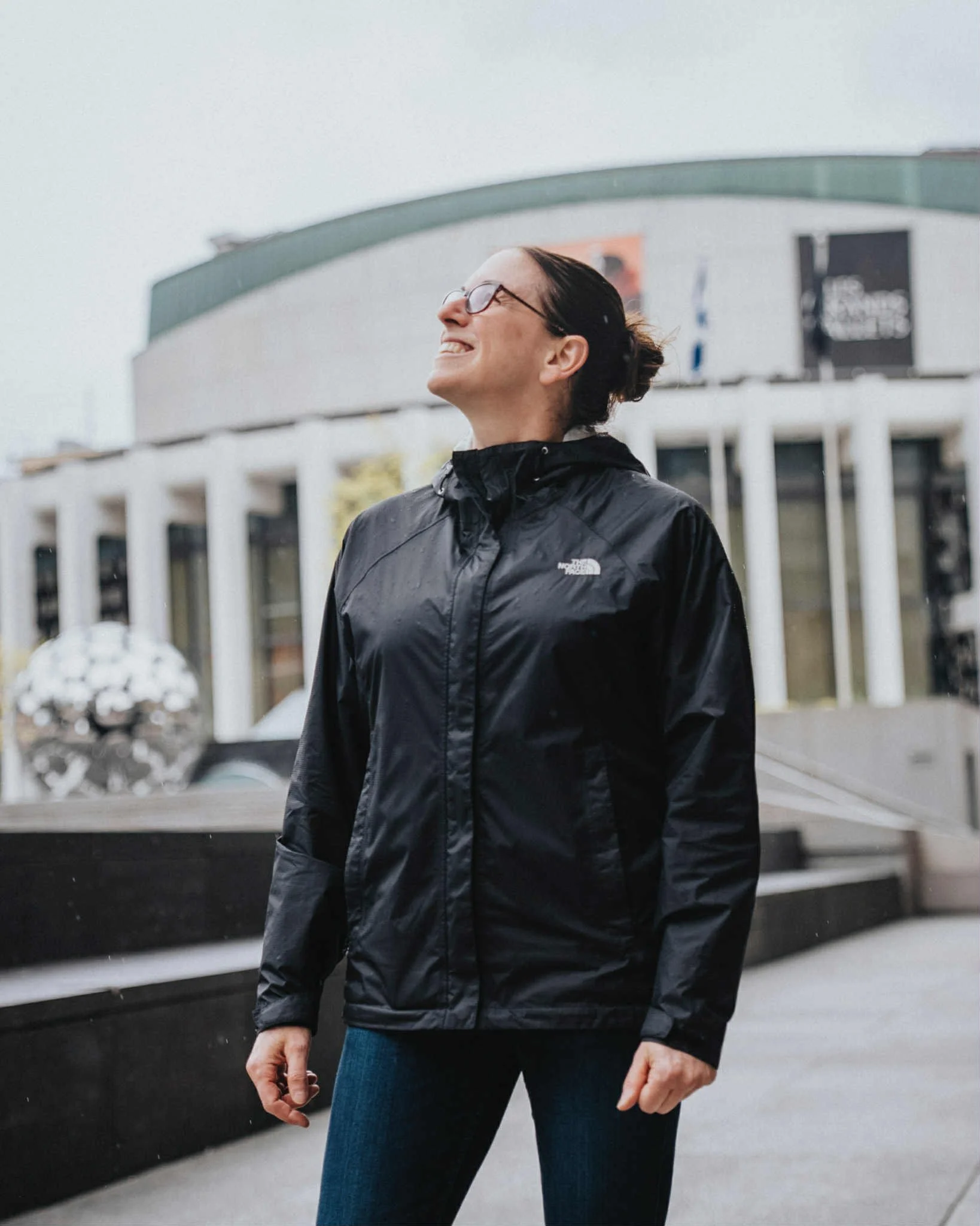 Femme souriante portant une veste imperméable noire de North Face, debout en extérieur devant un bâtiment moderne.