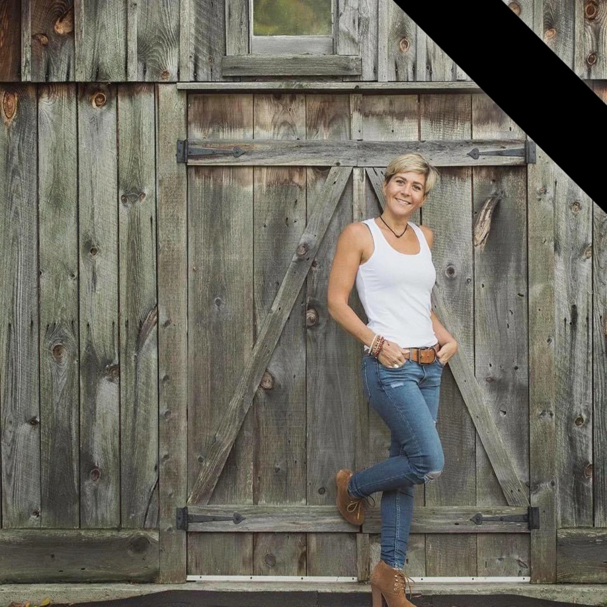 Une femme souriante debout devant une grande porte en bois antique, portant un débardeur blanc, un jean usé, des chaussures à talons marron, et quelques bracelets et un collier.