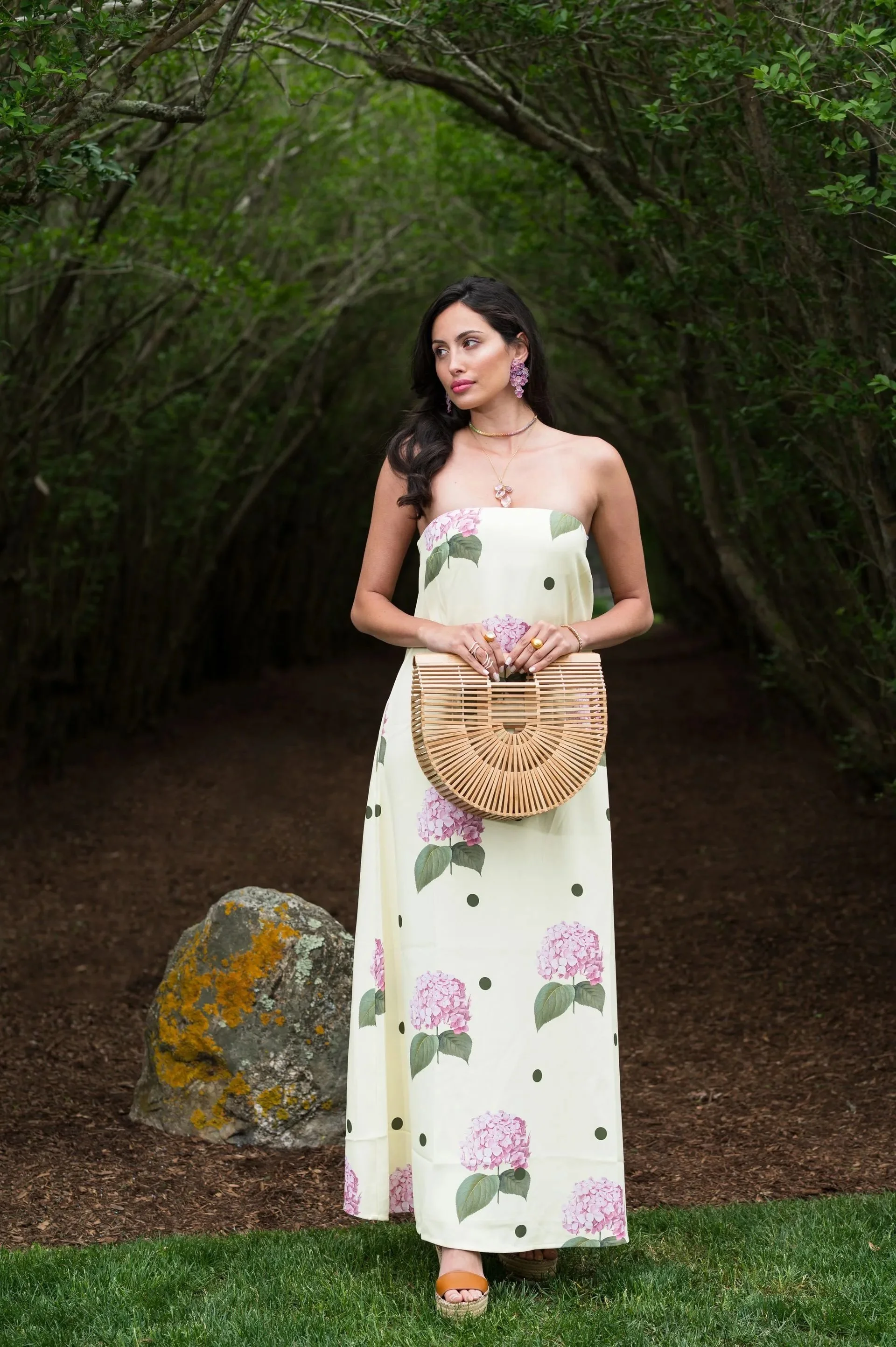 A woman in a strapless floral dress with pink hydrangea and green leaves print, standing outdoors on grass with a wooded background, holding a rattan clutch bag.