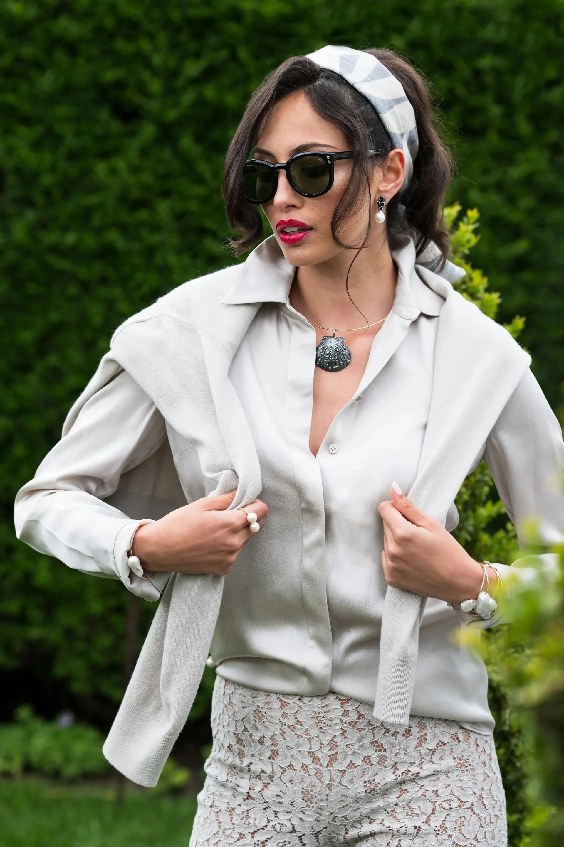 A stylish woman with dark hair, wearing large black sunglasses, a white blouse, a light cardigan draped over her shoulders, a patterned headscarf, and lace pants, stands outdoors with green foliage in the background.